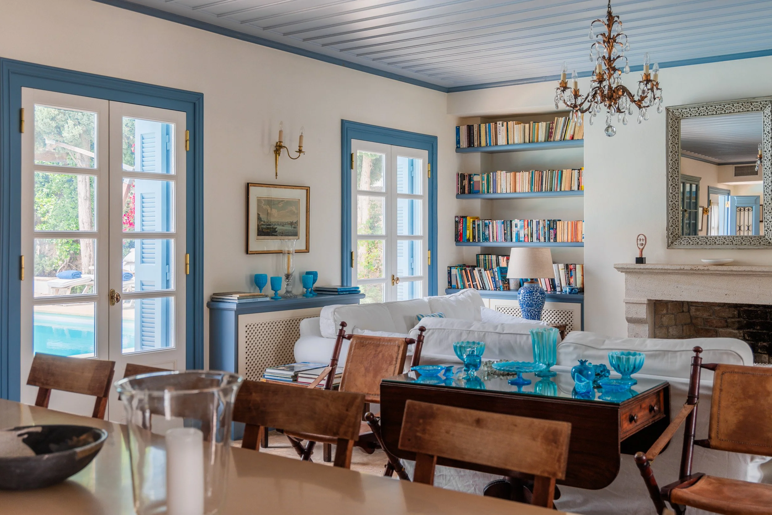 Living room with white sofas, wooden chairs, a glass-top table with blue glassware, a built-in bookshelf filled with books, a fireplace with a mirror above, and a chandelier hanging from the ceiling.