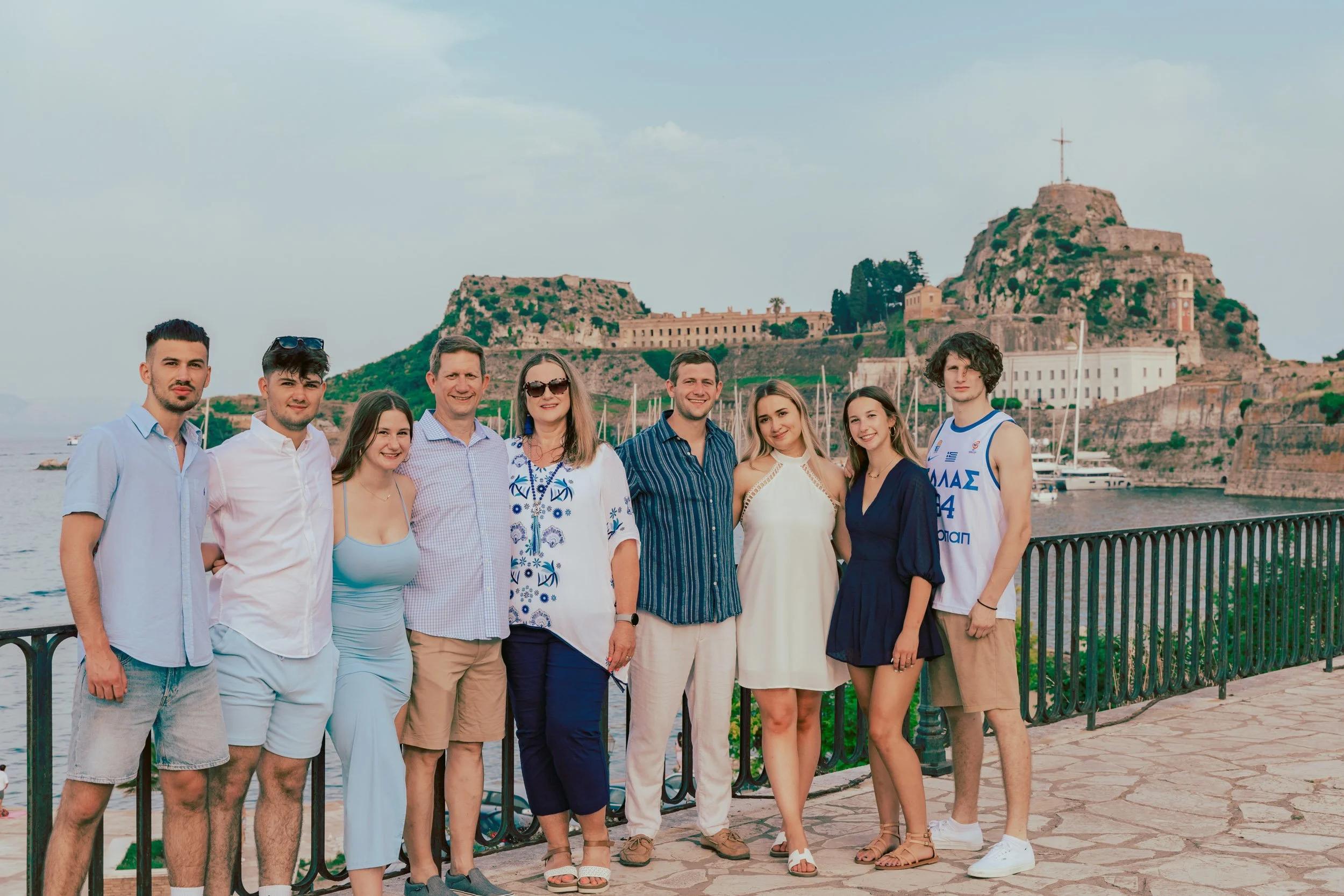 A group of nine people standing together on a waterfront promenade with a historic fortress and boats in the background.