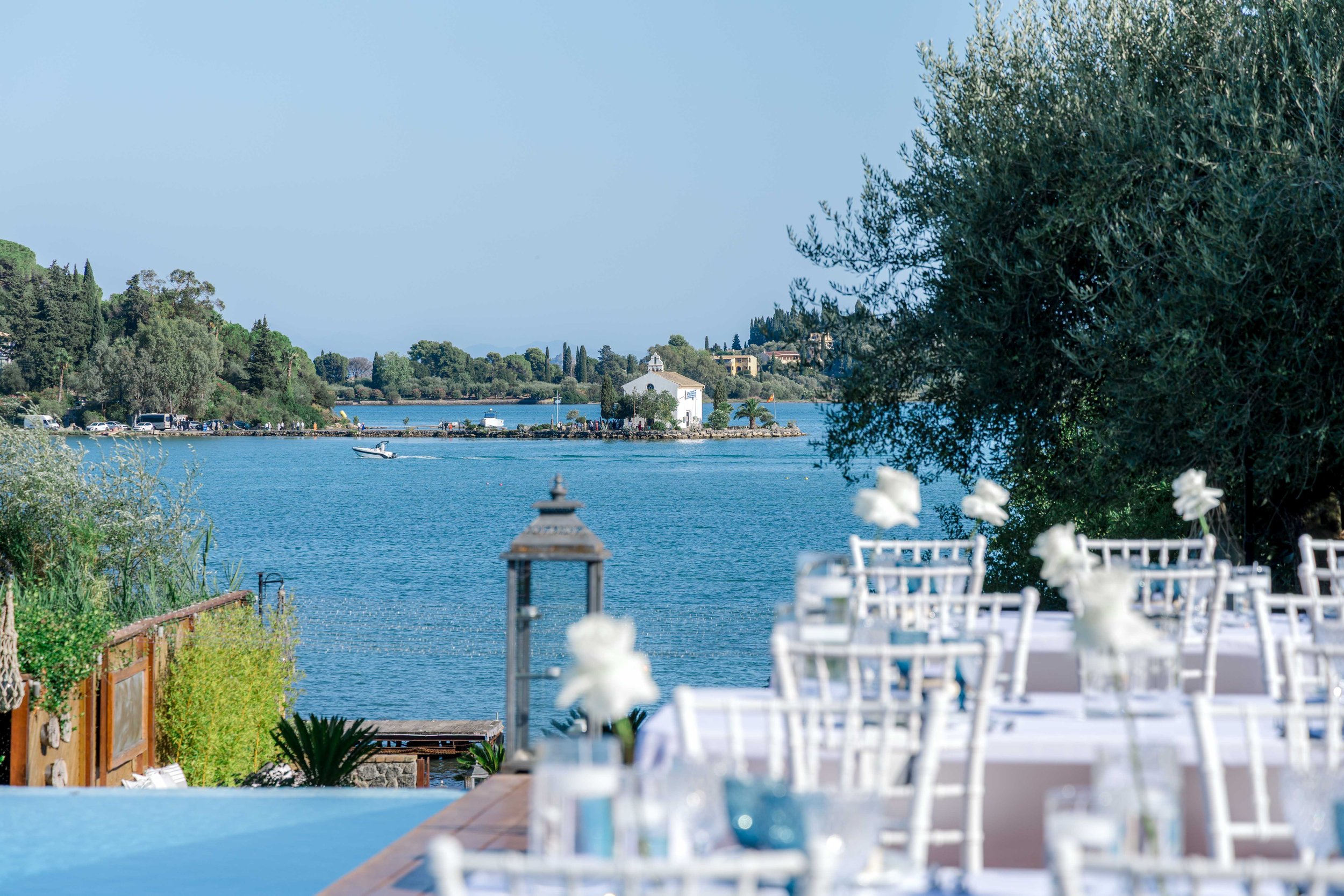 A lakeside outdoor dining area with white chairs and tablecloths, decorated with white flowers, overlooking a blue lake with a small island with a white building and trees in the background.