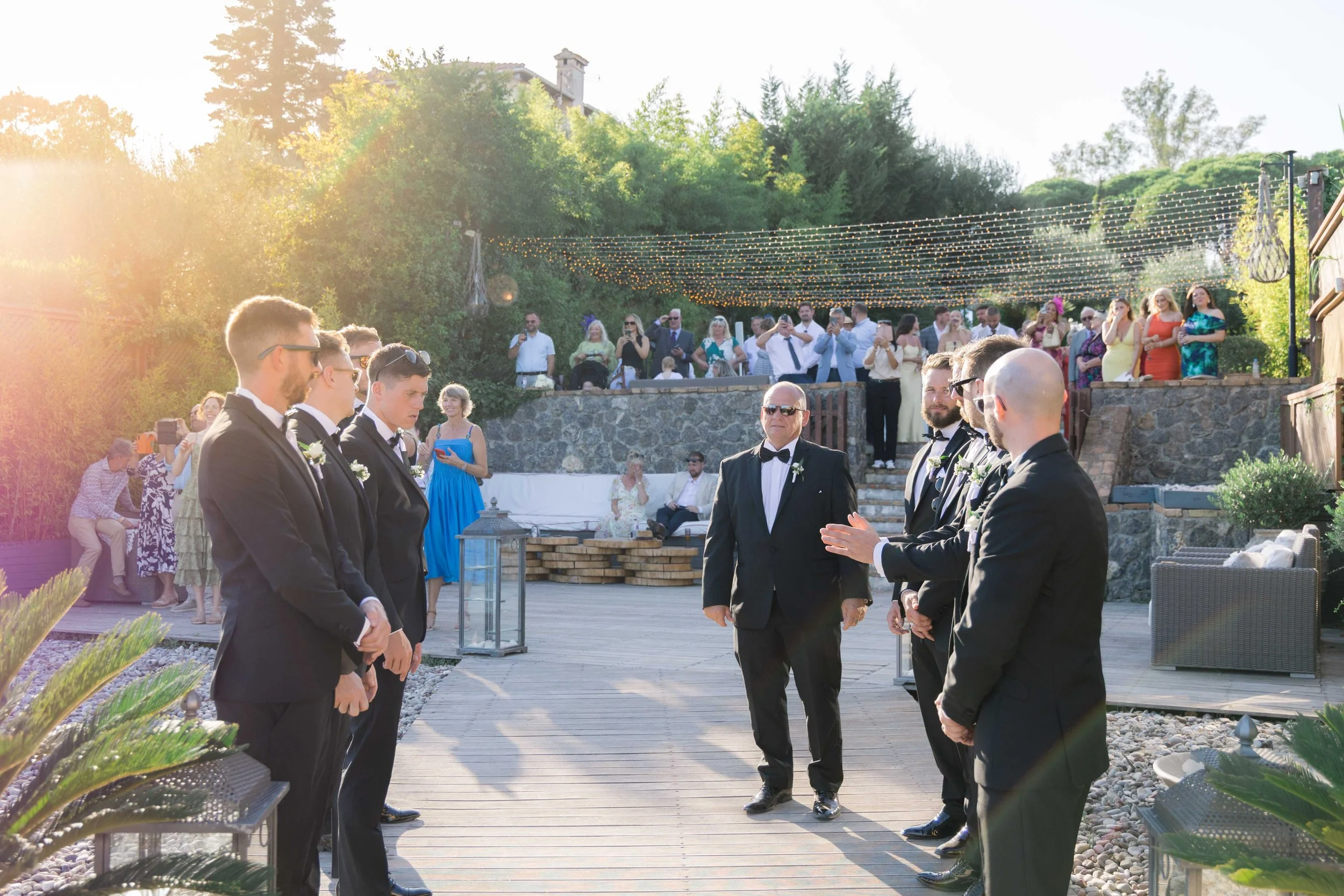 Men in black tuxedos standing in a line outdoors, facing a man in a tuxedo, during a wedding ceremony, with guests seated and standing behind, decorated with string lights and lush greenery.