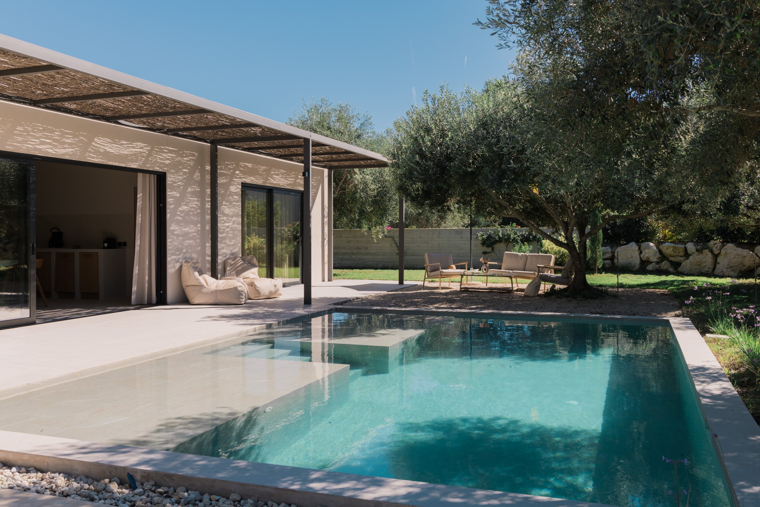 Modern backyard with a swimming pool, outdoor seating area with sofas, trees, and a house with an open patio under a sunny sky.
