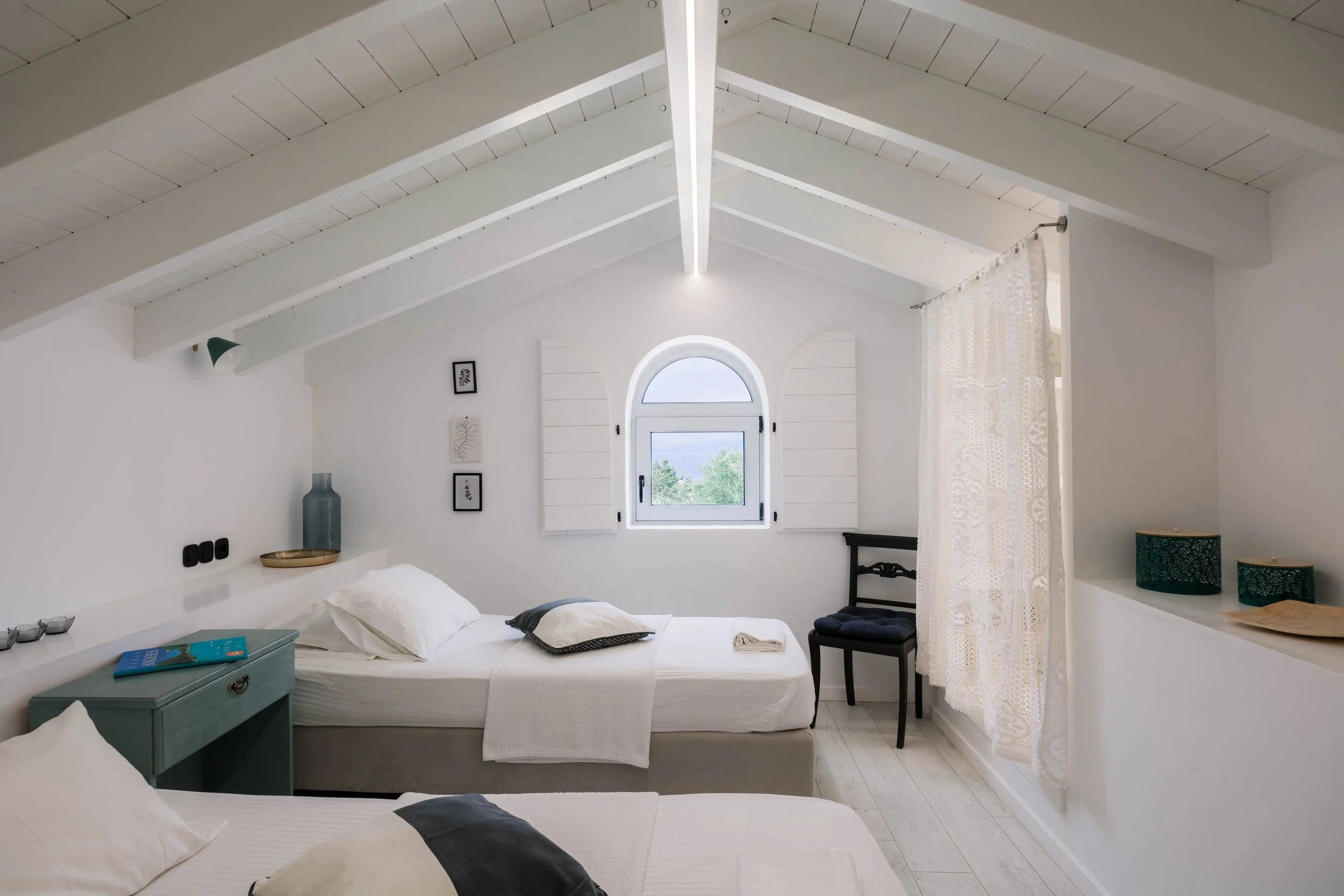 A bright, white minimalist bedroom with sloped ceiling, arched window with shutters, two beds, and decor in neutral tones.