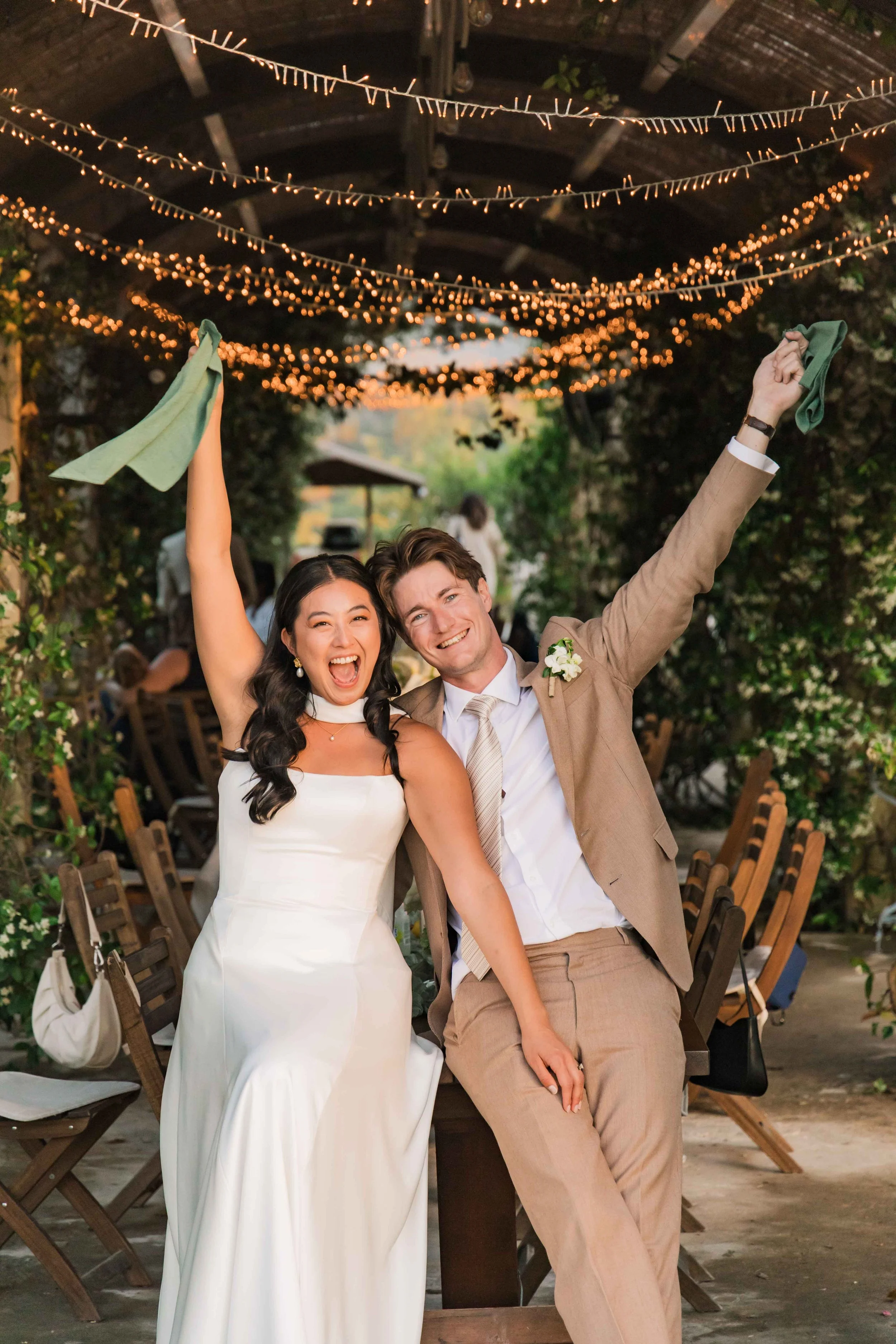 A newly married couple celebrating at their wedding reception outdoors beneath string lights. The bride is wearing a white wedding dress and the groom is in a tan suit. Both are smiling, with their arms raised, holding green napkins.