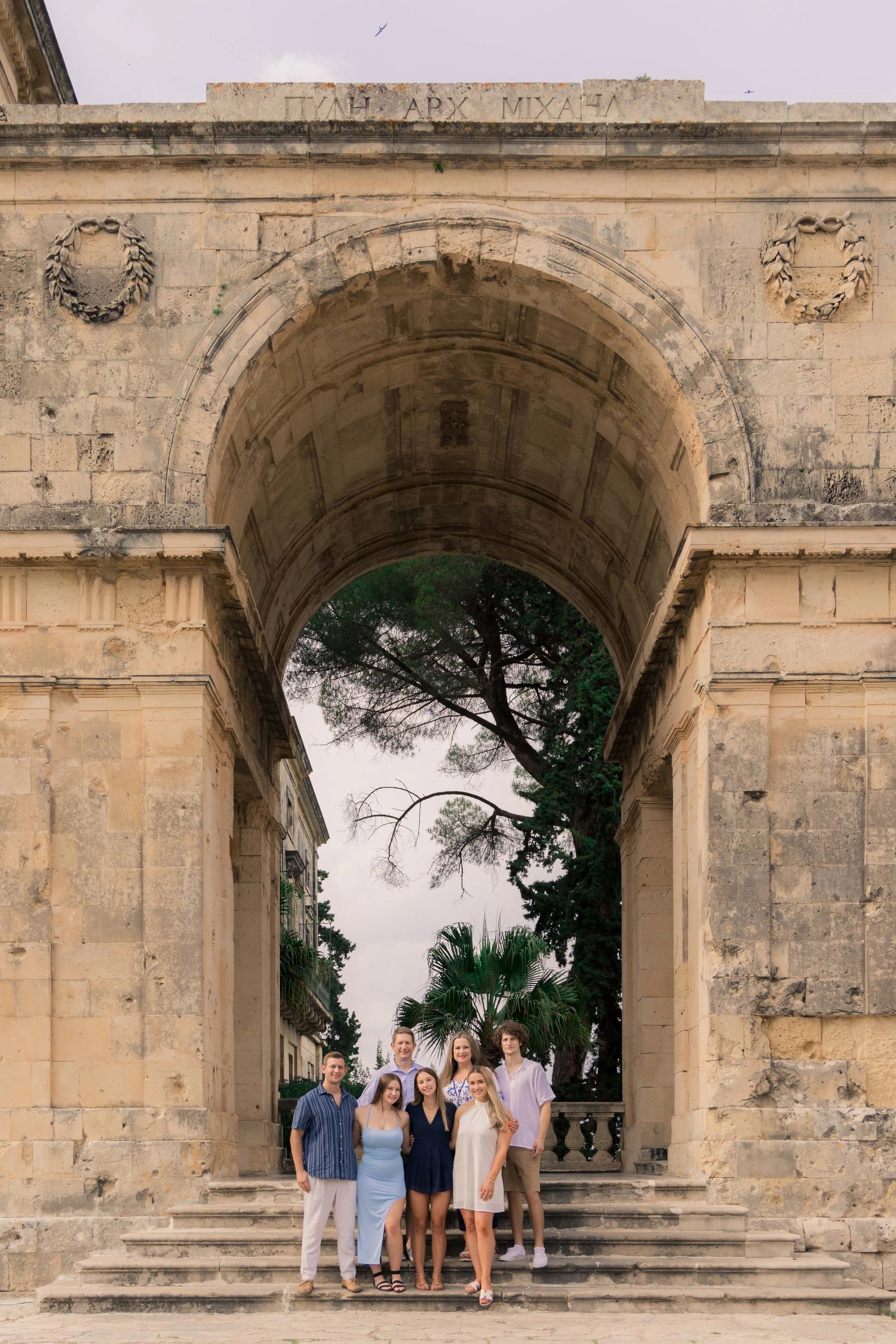 A group of seven young adults posing on steps under a historic stone archway at sunset, with trees visible in the background.