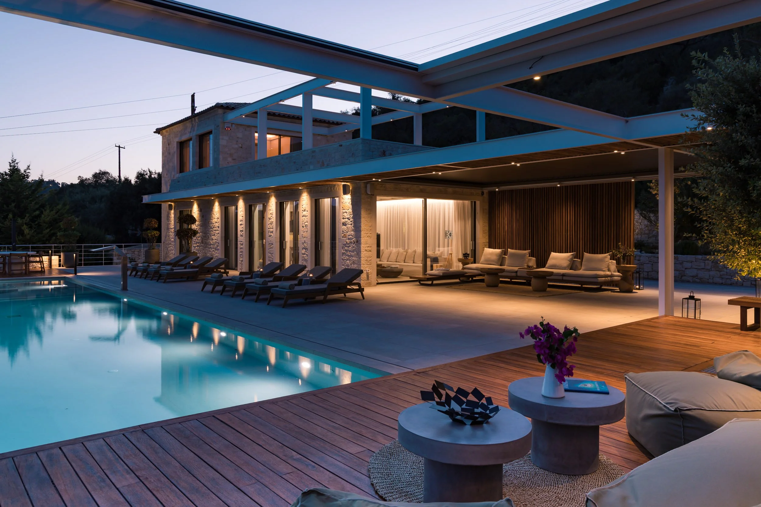 Modern two-story house with large outdoor patio and swimming pool at dusk, with lounge chairs, seating area, and decorative lighting.