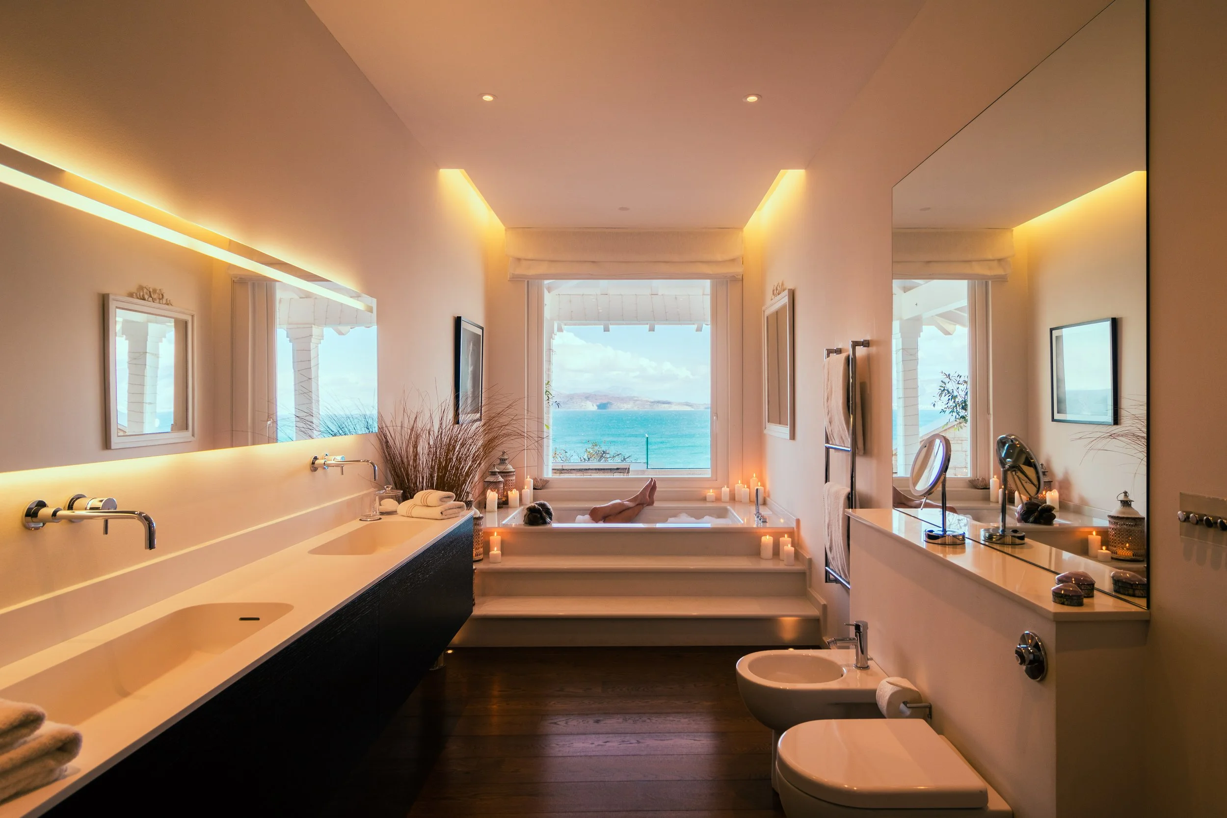 A luxurious bathroom with a view of the ocean, featuring a large bathtub with candles and a person relaxing in it, a double sink vanity with a mirror, and a bidet; the room is illuminated with warm lighting.