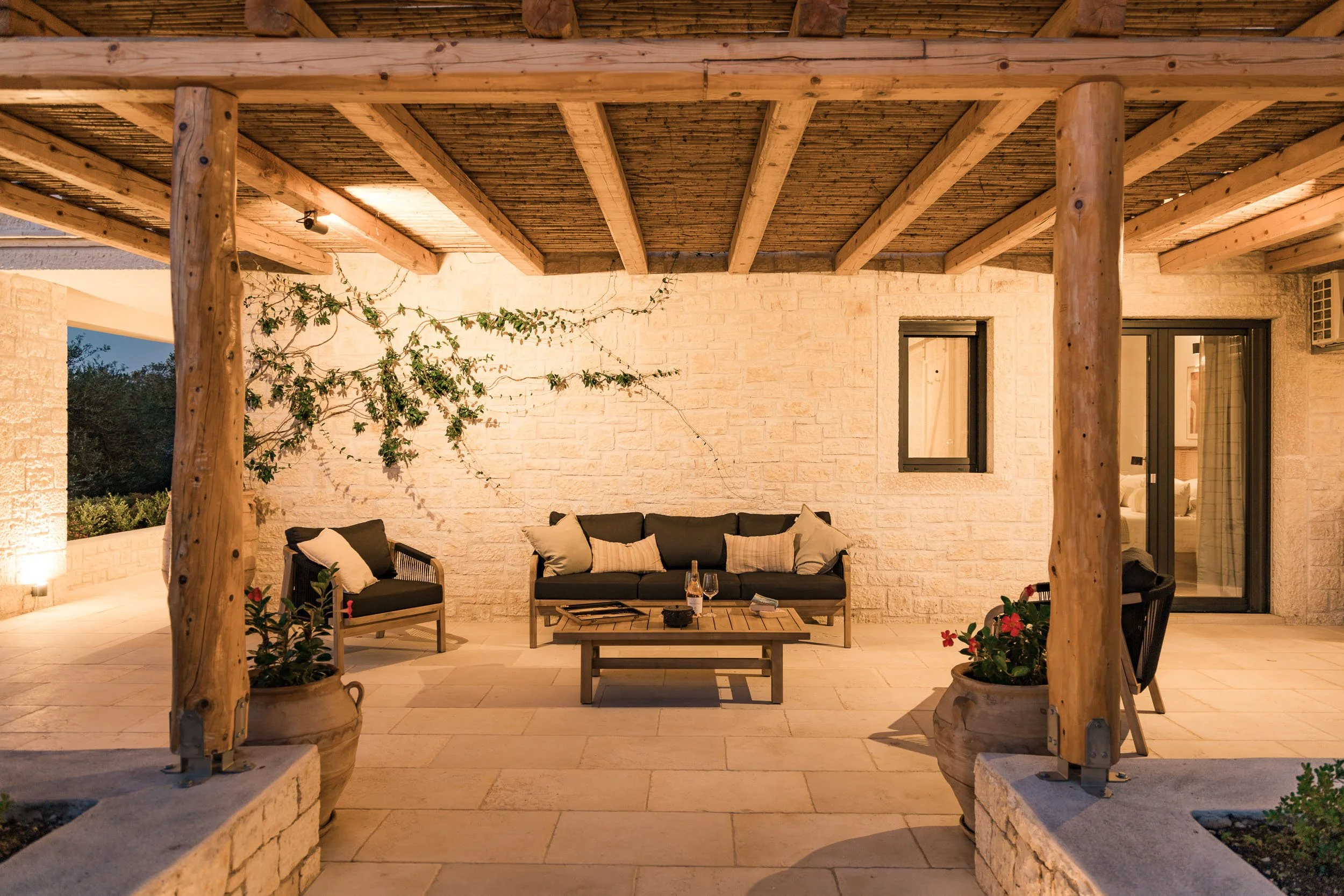 Under a wooden pergola, outdoor patio with black cushioned seating, throw pillows, coffee table with drinks, potted plants, and a brick wall in the background.