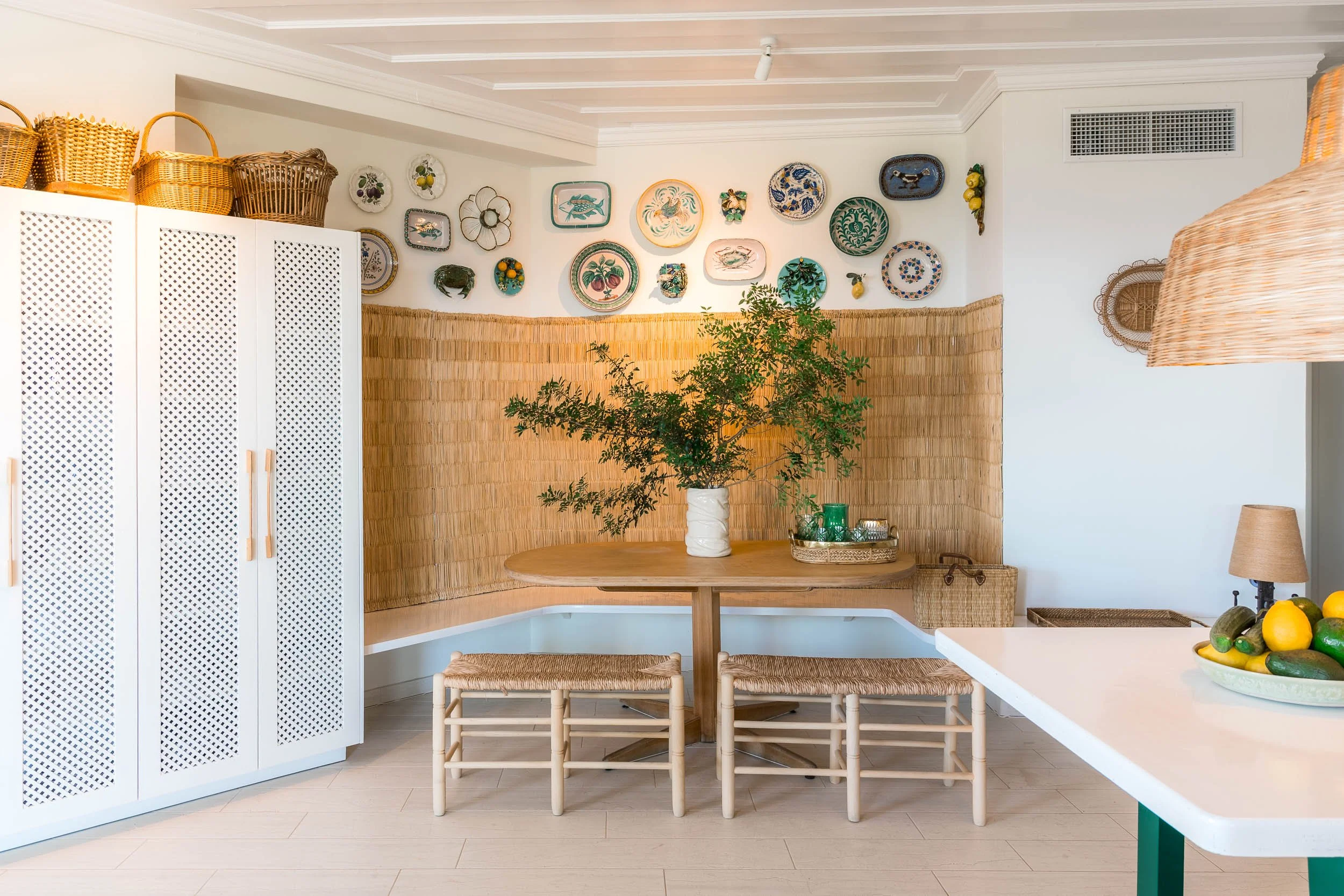 A cozy dining nook with a wooden round table, surrounded by four woven stools, placed against a bamboo wall. The table features a large vase with green foliage and a tray with candles. The wall above is decorated with colorful plates and wall art. To
