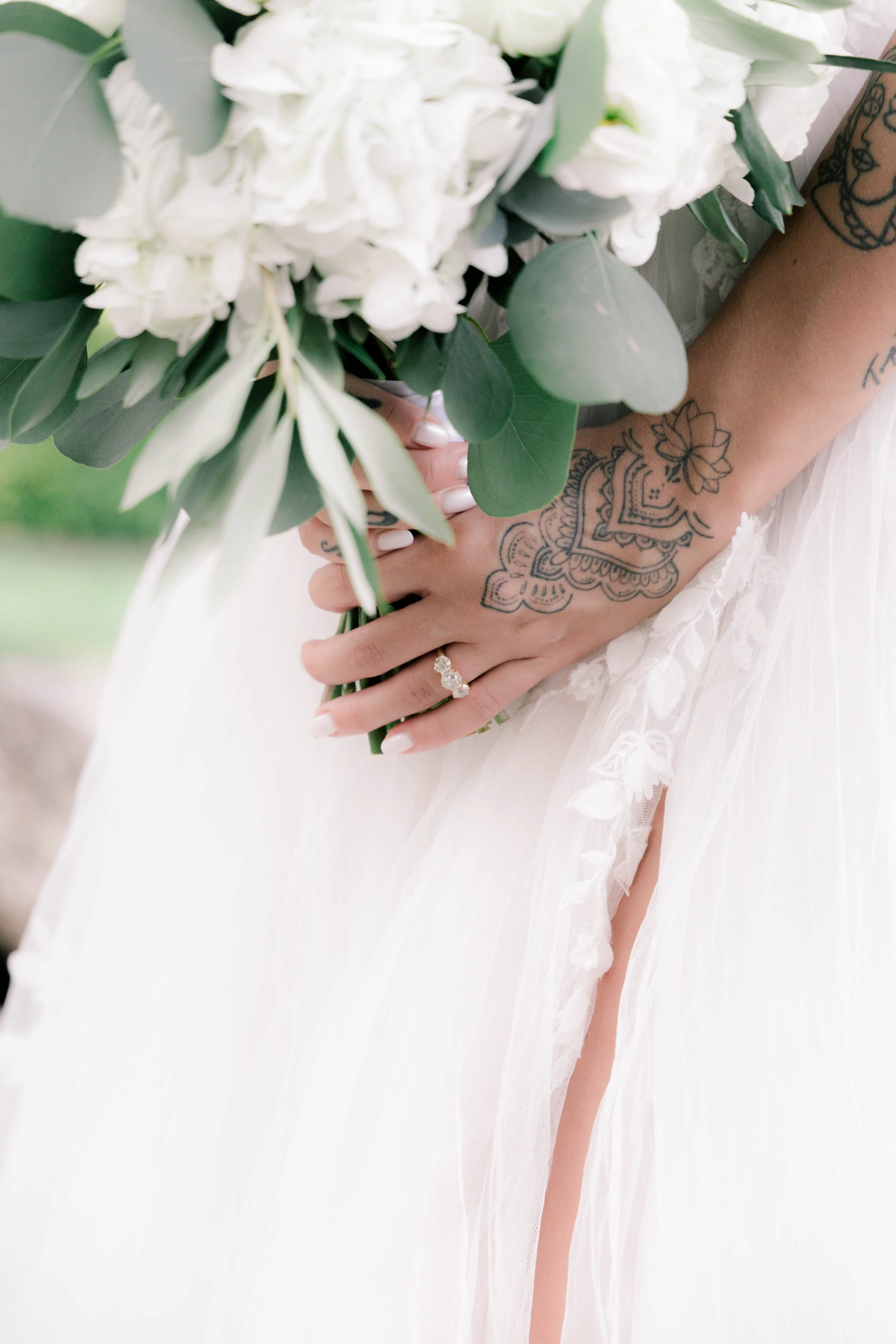 A bride holding a bouquet of white flowers and green leaves, showing her hand with an engagement ring and a colorful tattoo, wearing a white wedding dress.