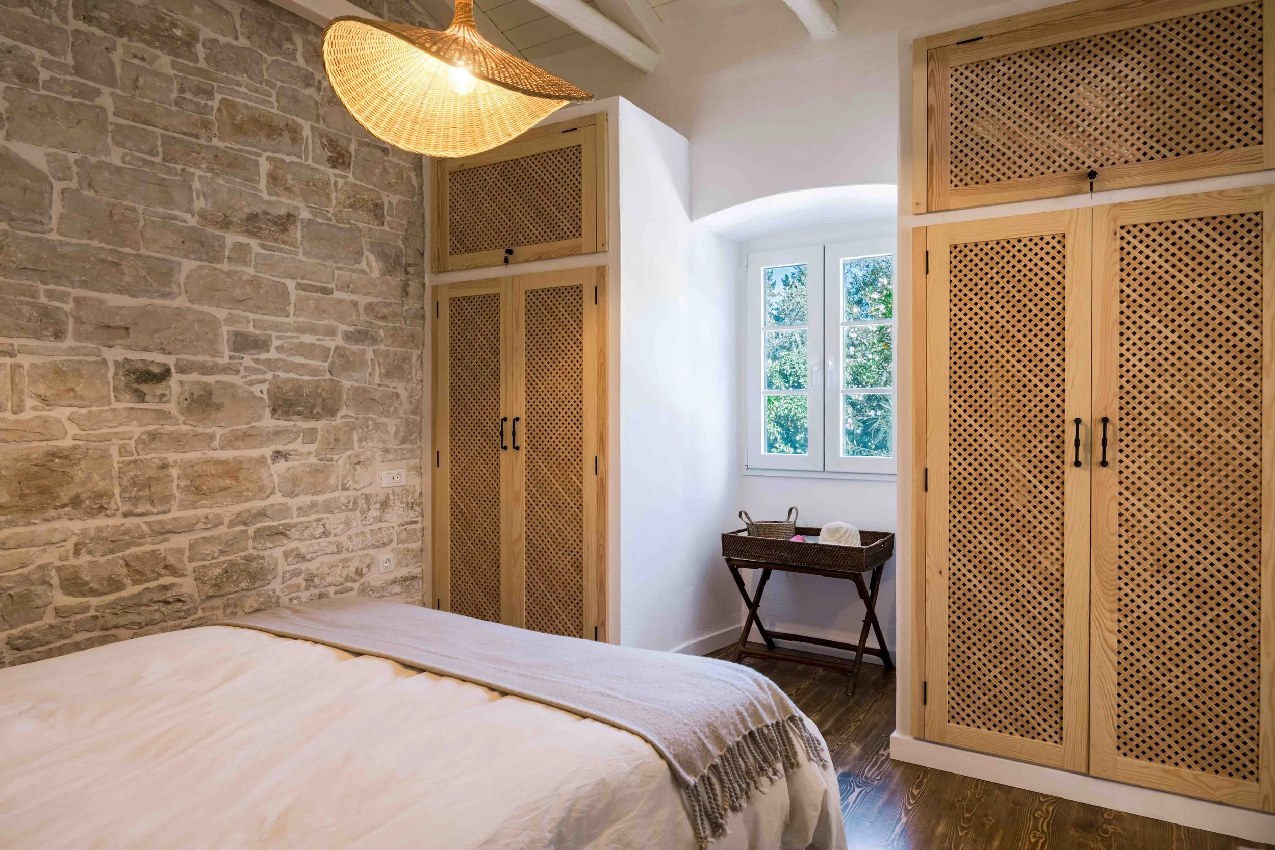A cozy bedroom with a brick accent wall, wooden cabinets with lattice doors, a small window with green trees outside, and a bed with white linens and a beige throw blanket.