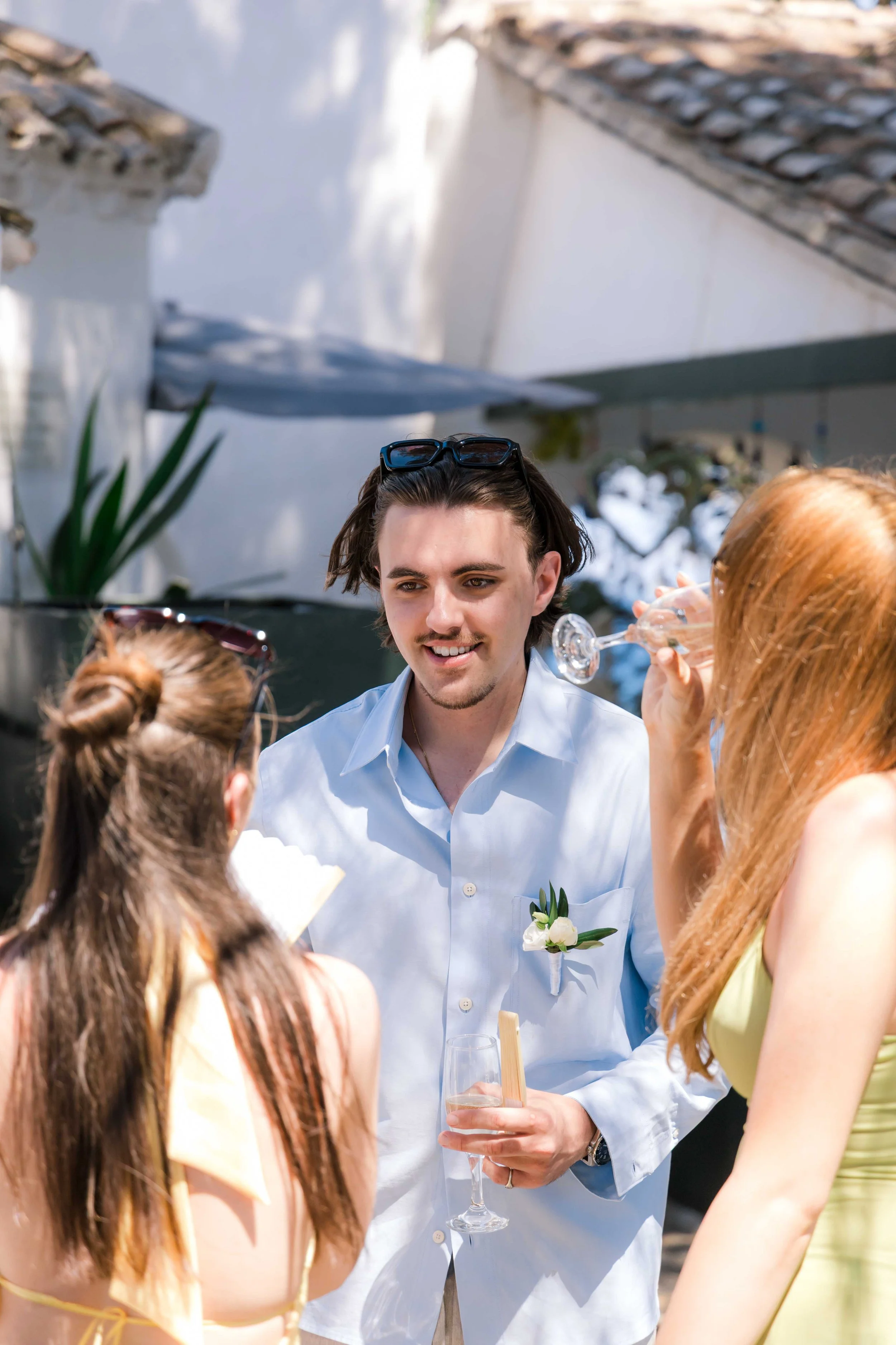 A young man with dark hair wearing a light blue shirt with a white boutonniere, holding a glass of champagne, talking to two women at an outdoor event.