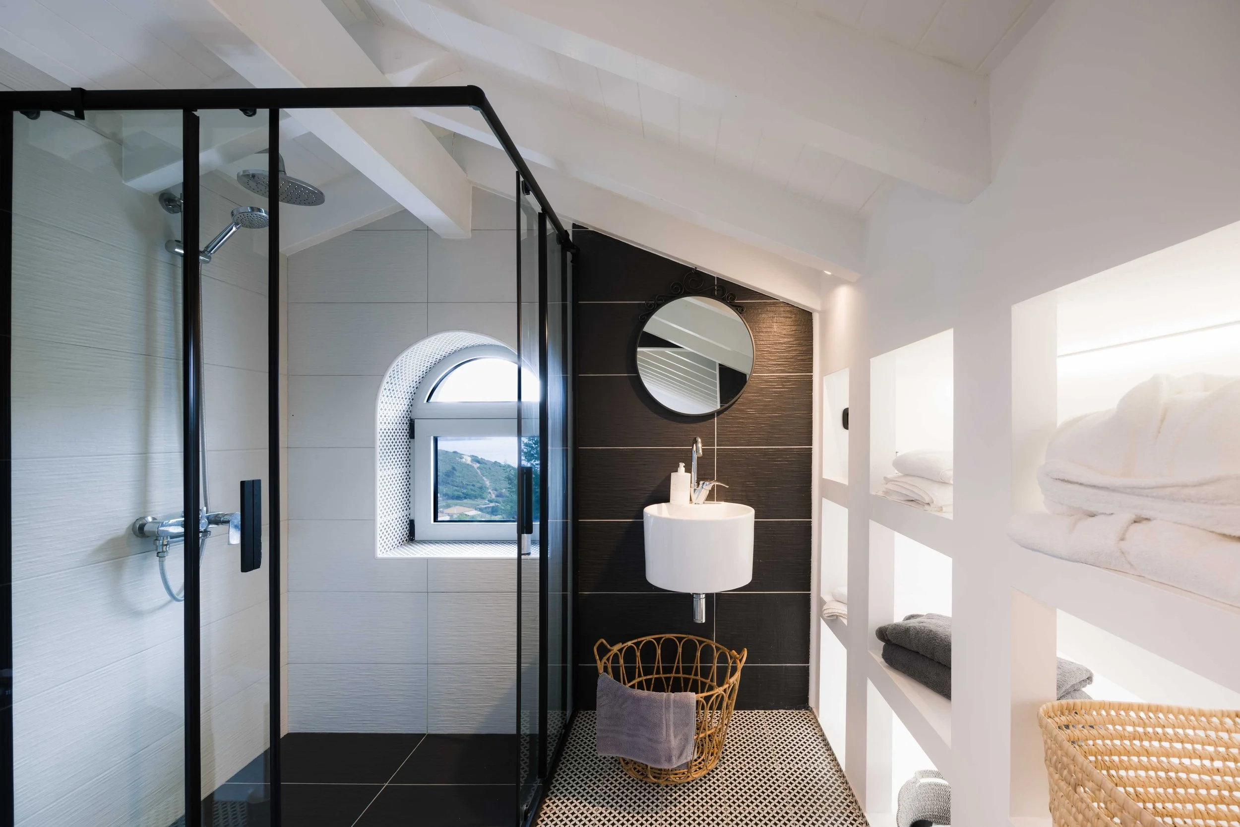 Modern bathroom with glass-enclosed shower, arched window, round mirror, white wall-mounted sink, wicker laundry basket, and built-in shelves with folded towels.