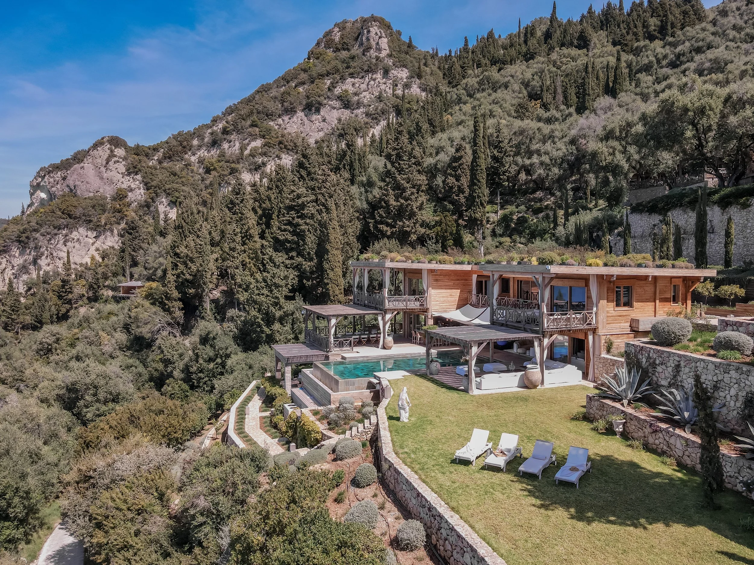 A modern wooden house with multiple terraces and a swimming pool, situated on a hillside surrounded by greenery and mountains.