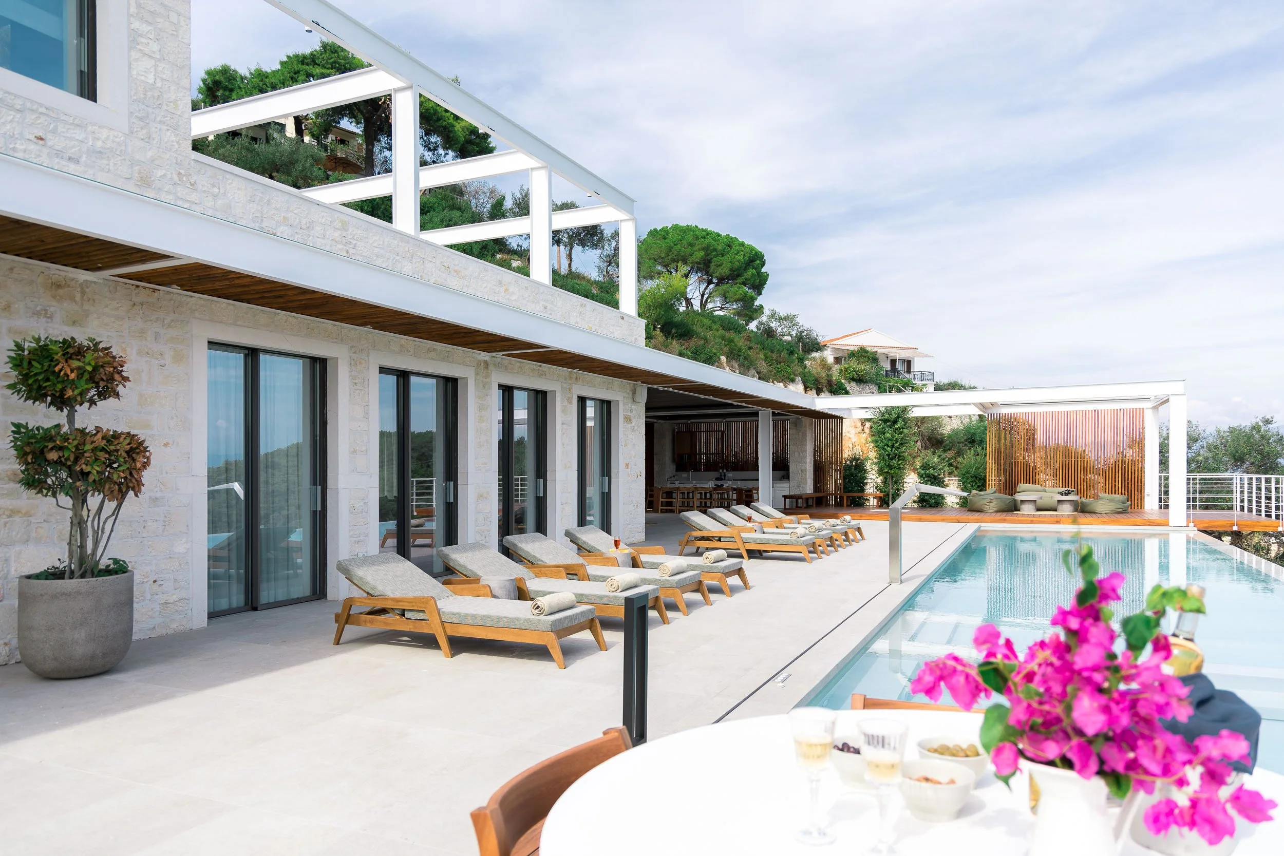 Luxury outdoor poolside lounge area with sunbeds, a dining table with drinks, and a view of hills and greenery.