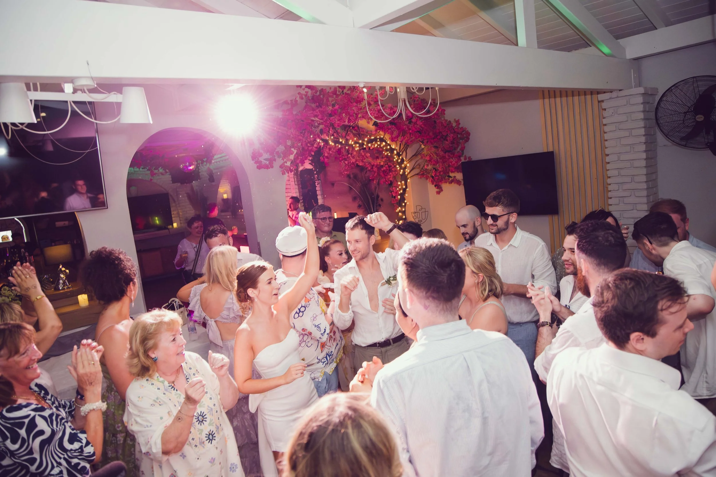 People dancing and celebrating at a lively indoor party or wedding reception with colorful lighting and decorations.