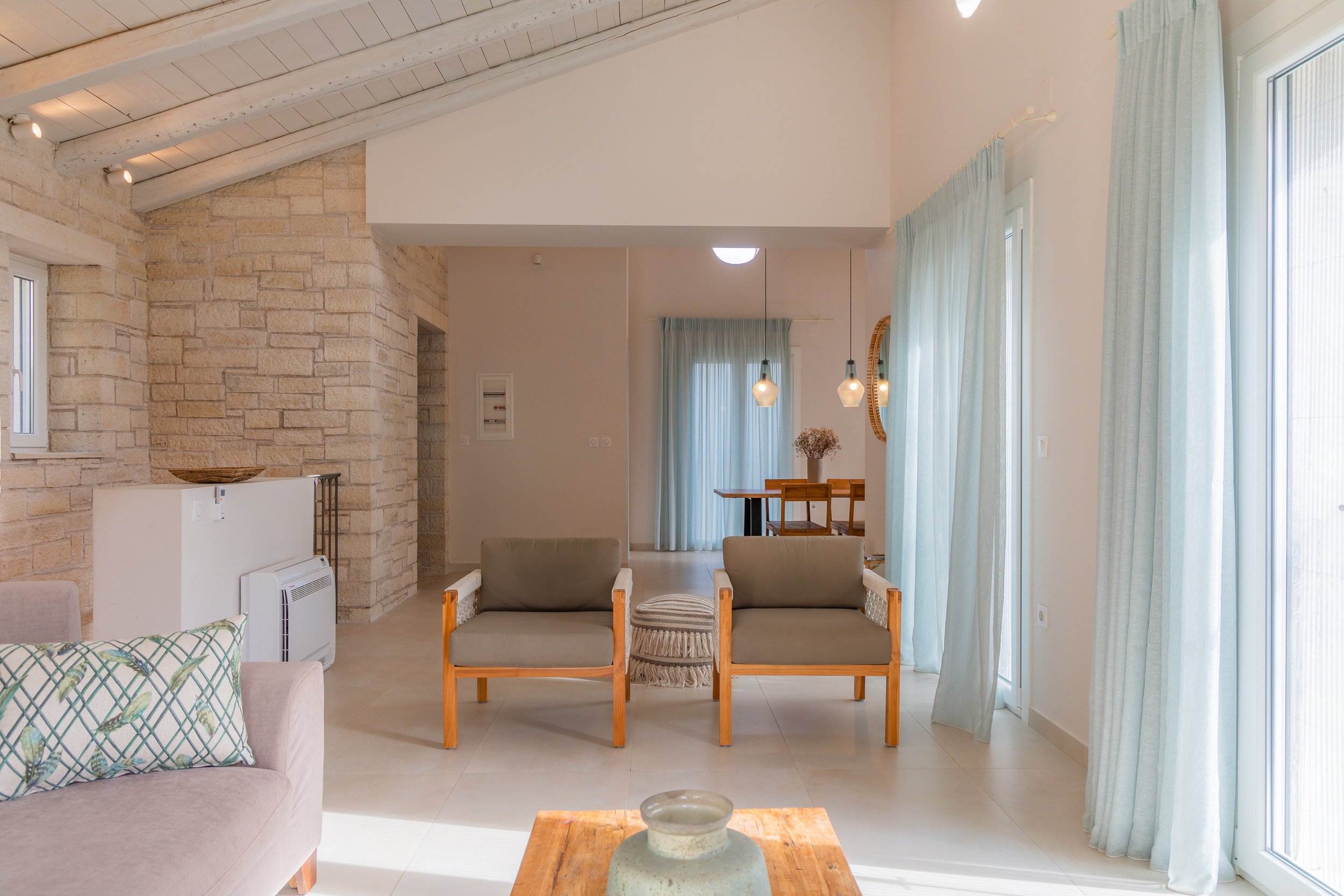 Bright living room with light-colored walls, large windows with sheer curtains, two wooden armchairs with gray cushions, a beige sofa with patterned pillows, a small wooden coffee table, and a dining area with a wooden table and chairs in the backgro