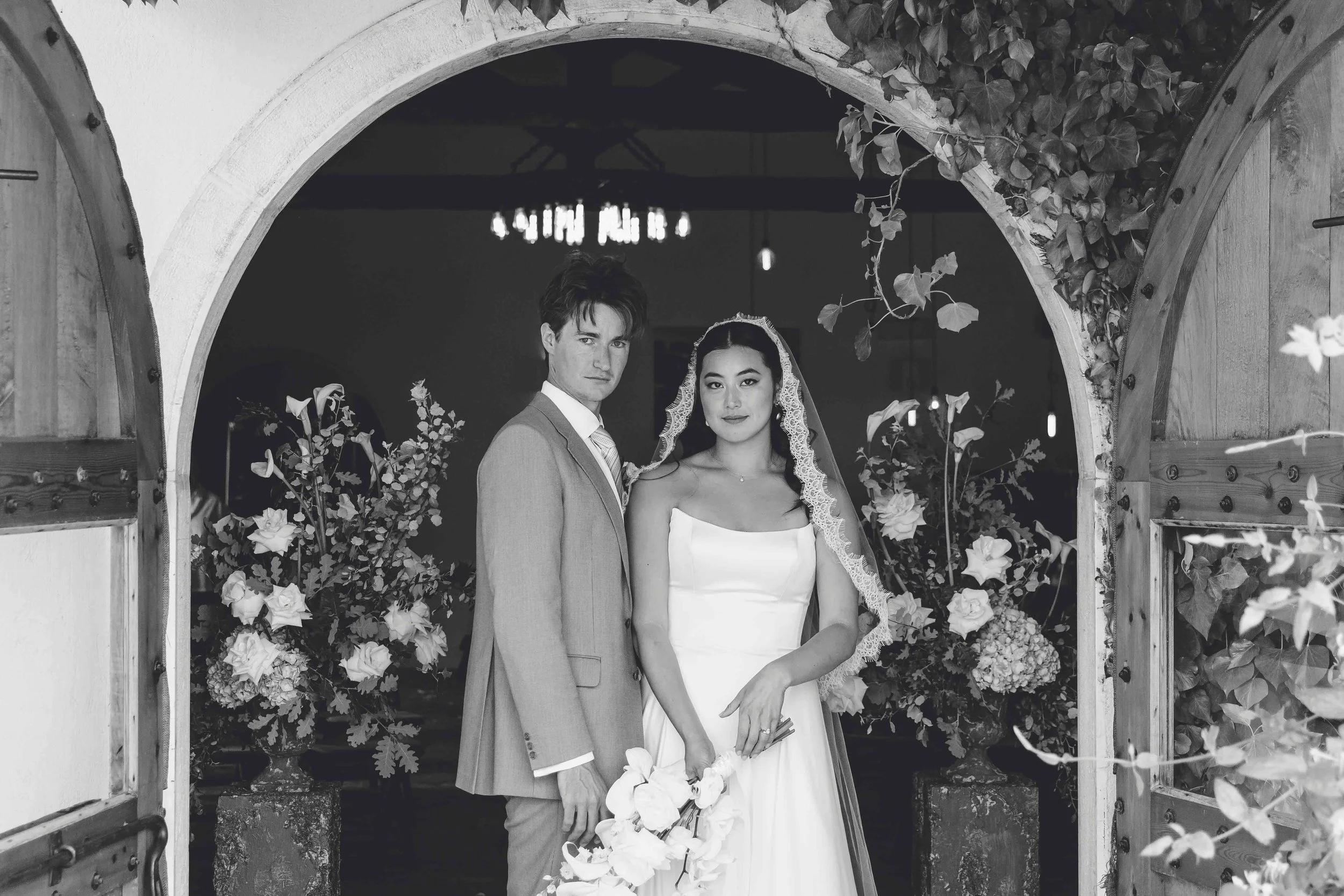 Black and white photo of a bride and groom standing under a wooden archway with flowers.
