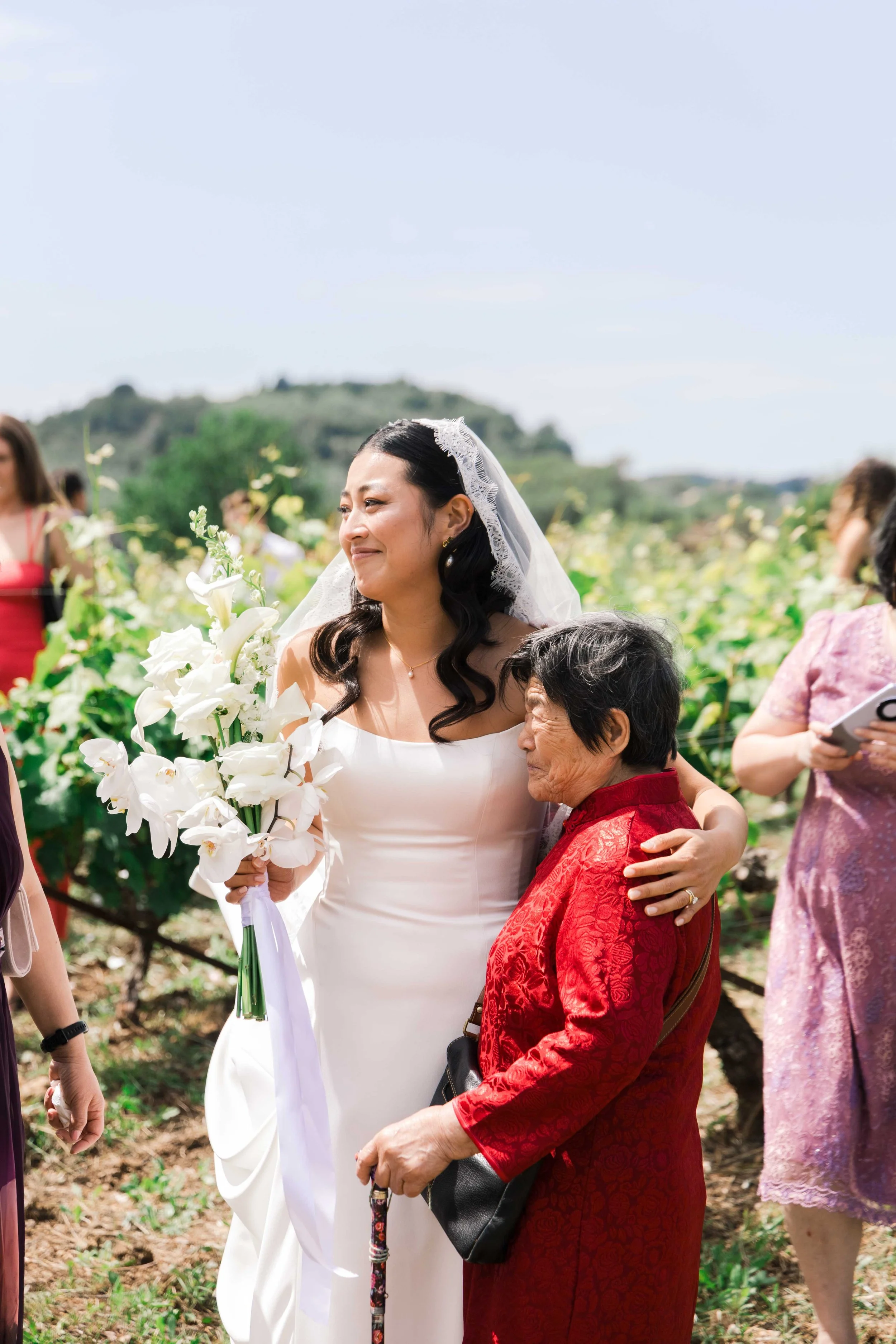 A bride in a white wedding dress holding a bouquet of white flowers, sharing a tender moment and hugging an elderly woman in a red dress outdoors during a sunny day, with other people and greenery in the background.