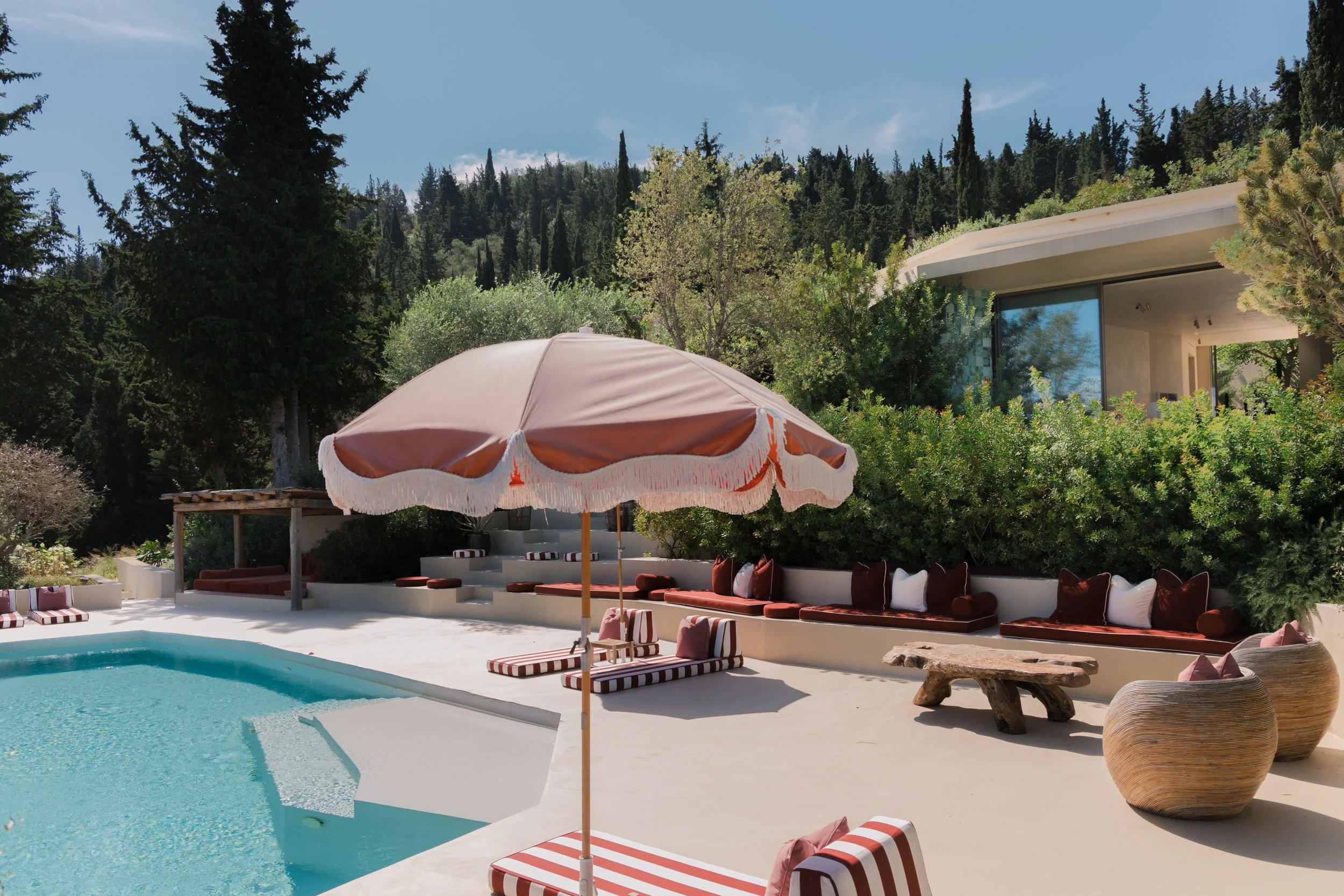 Luxury outdoor swimming pool area with poolside lounge chairs, striped umbrellas, and surrounding lush green trees and bushes, modern house with large glass windows in the background.