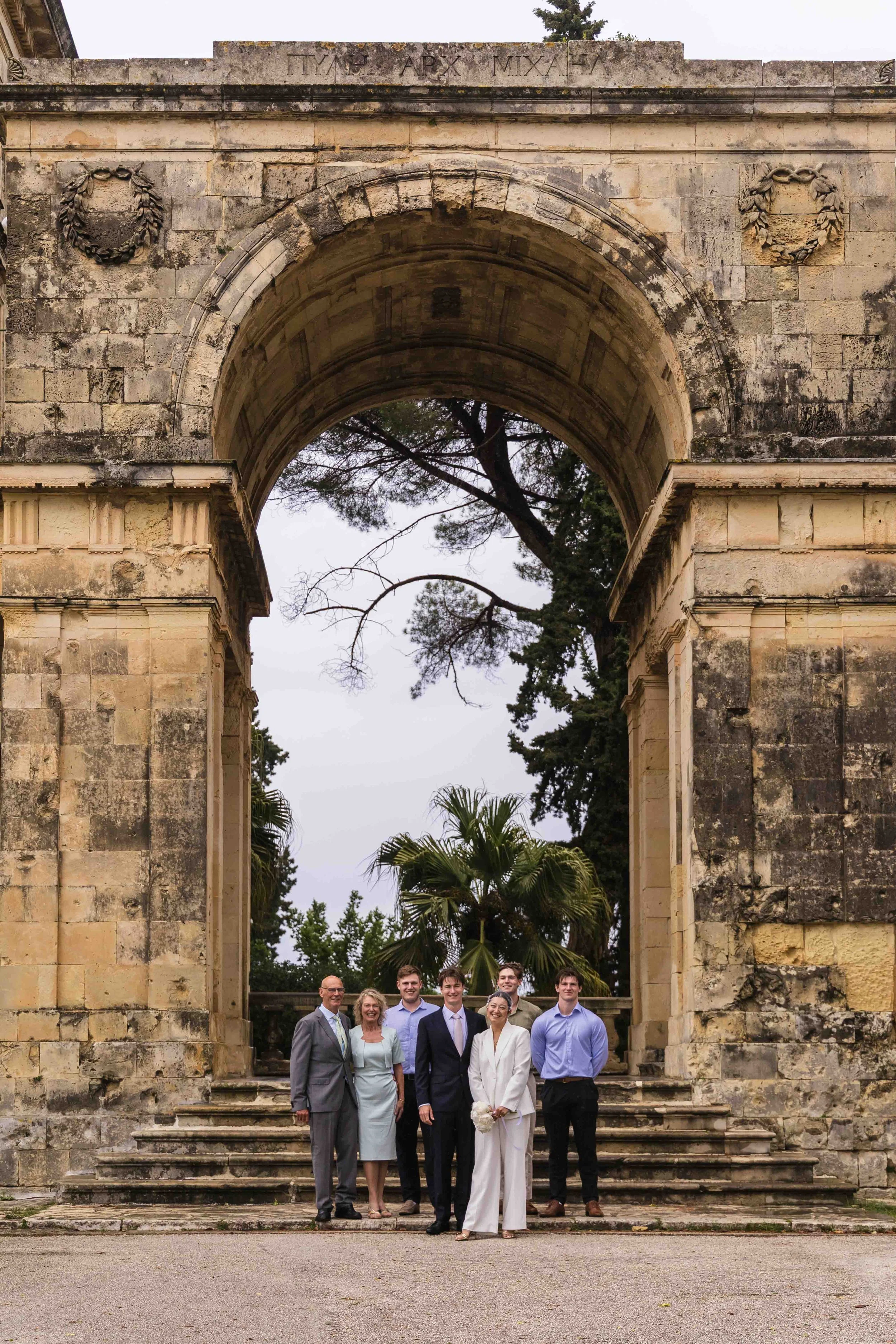 Group of seven people standing on steps in front of an ancient stone archway, with trees in the background, celebrating a special occasion.