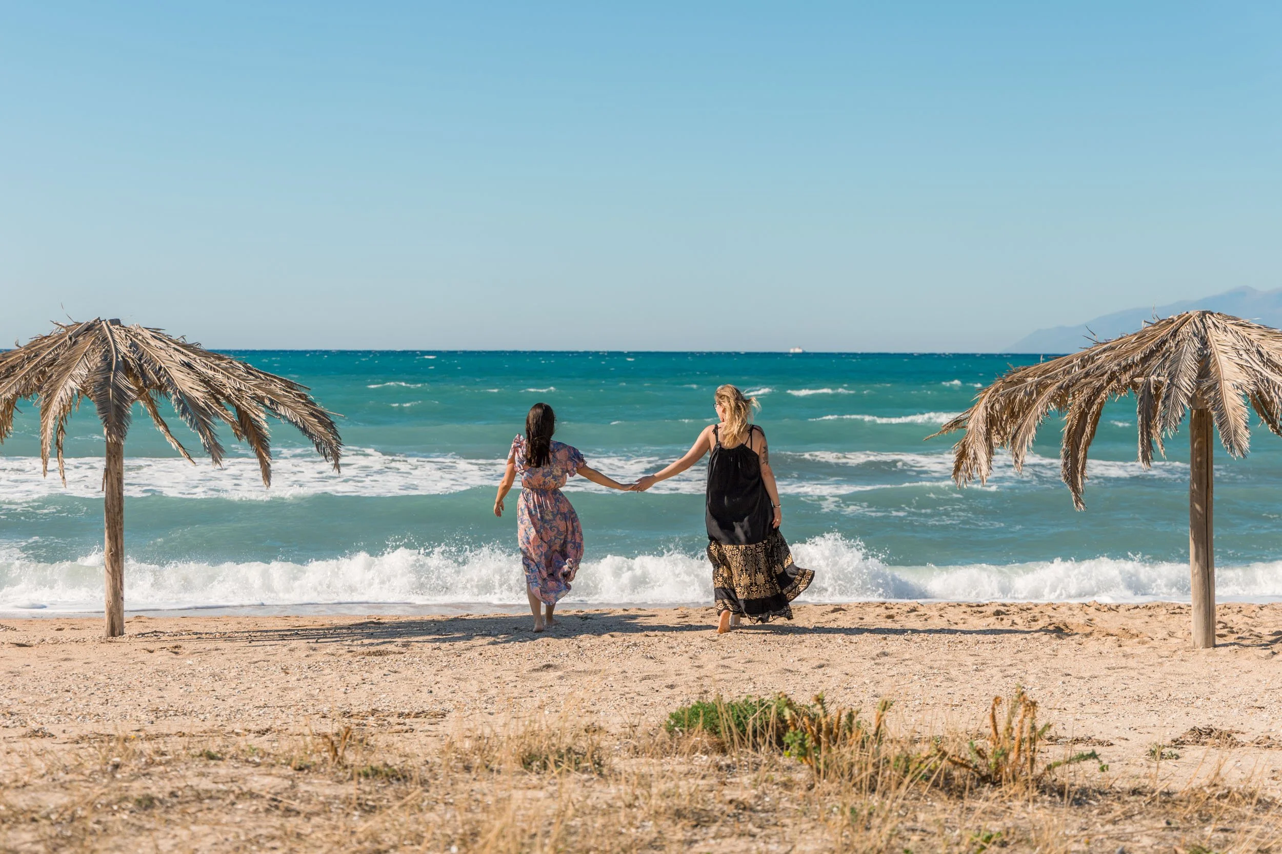 Two women walking hand in hand on the beach near the ocean, surrounded by straw umbrellas.