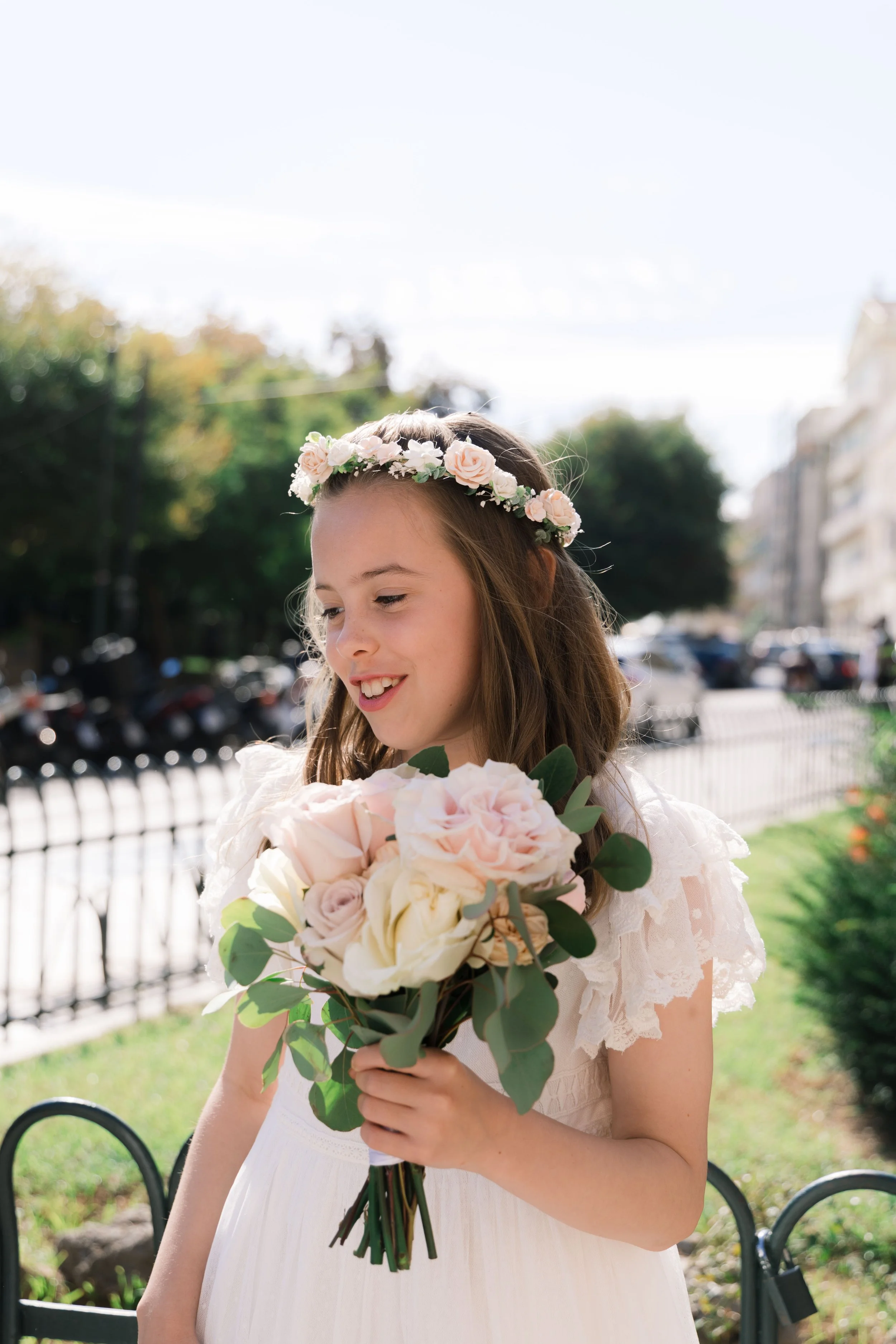 A young girl dressed in a white dress with lace sleeves holds a bouquet of pink and white roses with green leaves, standing outdoors in sunlight with a city street and trees in the background.