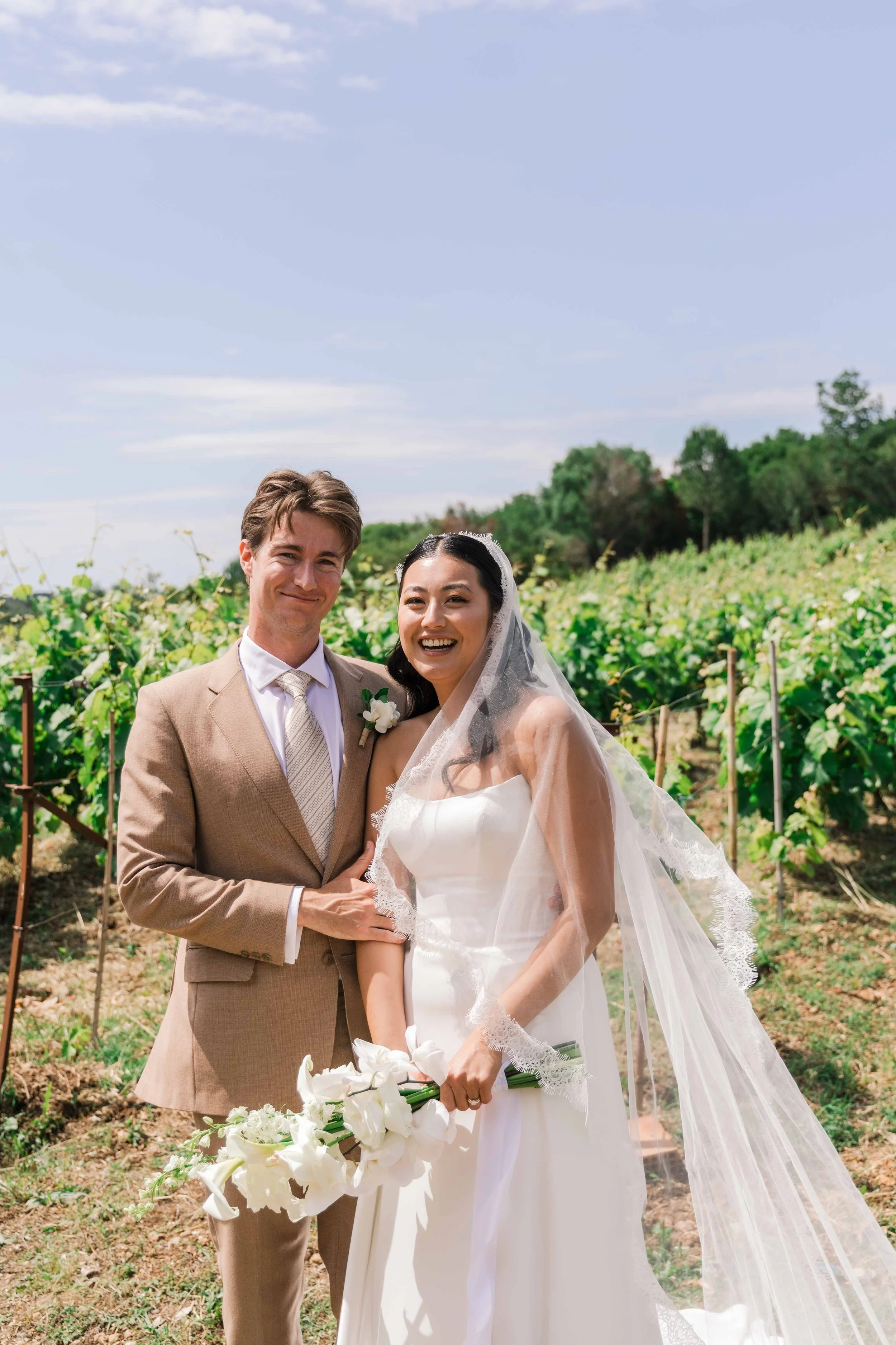 Happy bride and groom standing together outdoors on a sunny day, with greenery in the background, celebrating their wedding.