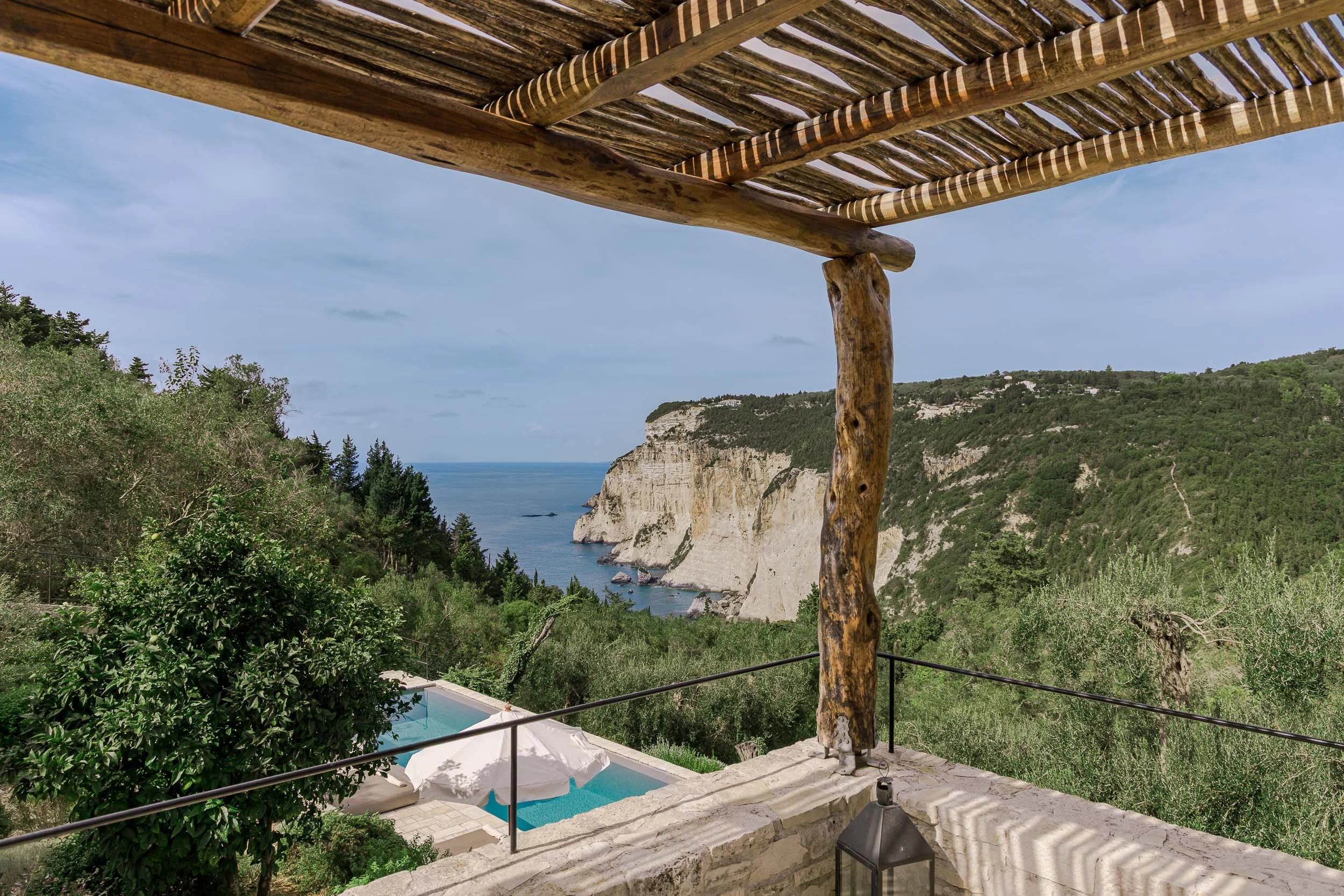 A scenic view of ocean cliffs with green trees, taken from a balcony with wooden beams and a railing, overlooking a swimming pool with an umbrella.