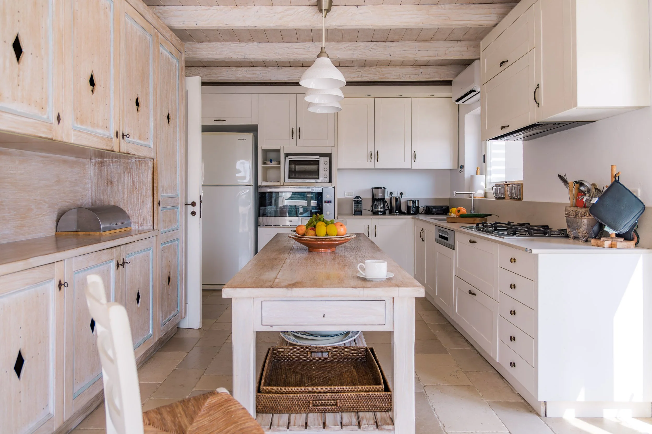 A kitchen with white cabinets, a wooden island, and a ceiling with wooden beams. The counter has various appliances and a bowl of fruit, and the room is well-lit with natural light.