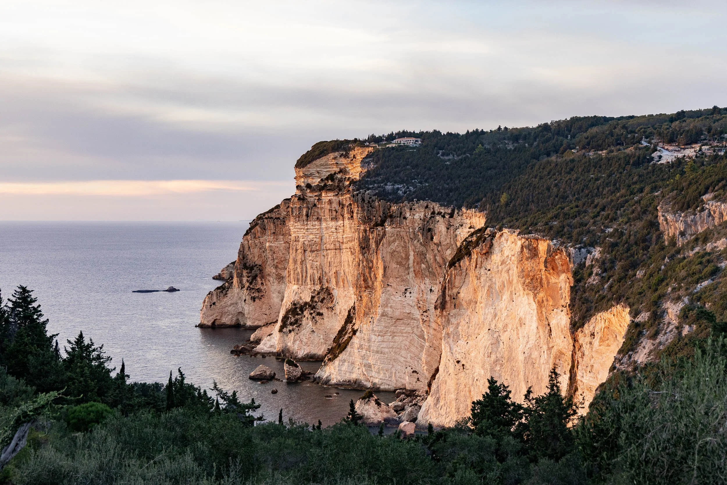 Cliffside view of Tall white rock formations along the coast with greenery in the foreground and hills in the background.