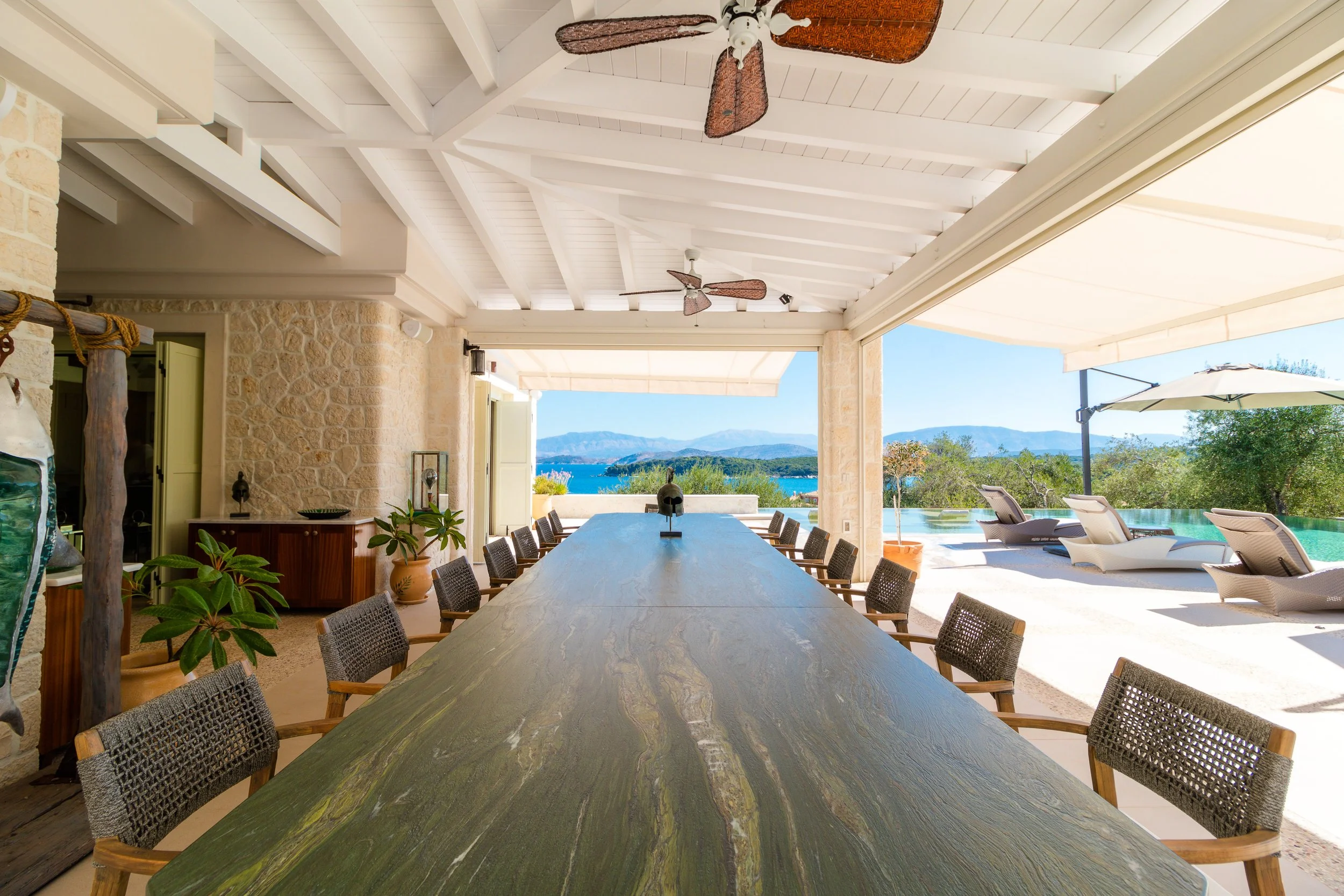 Outdoor dining area with a large wooden table, wicker chairs, potted plants, and a view of a pool and mountains in the background.