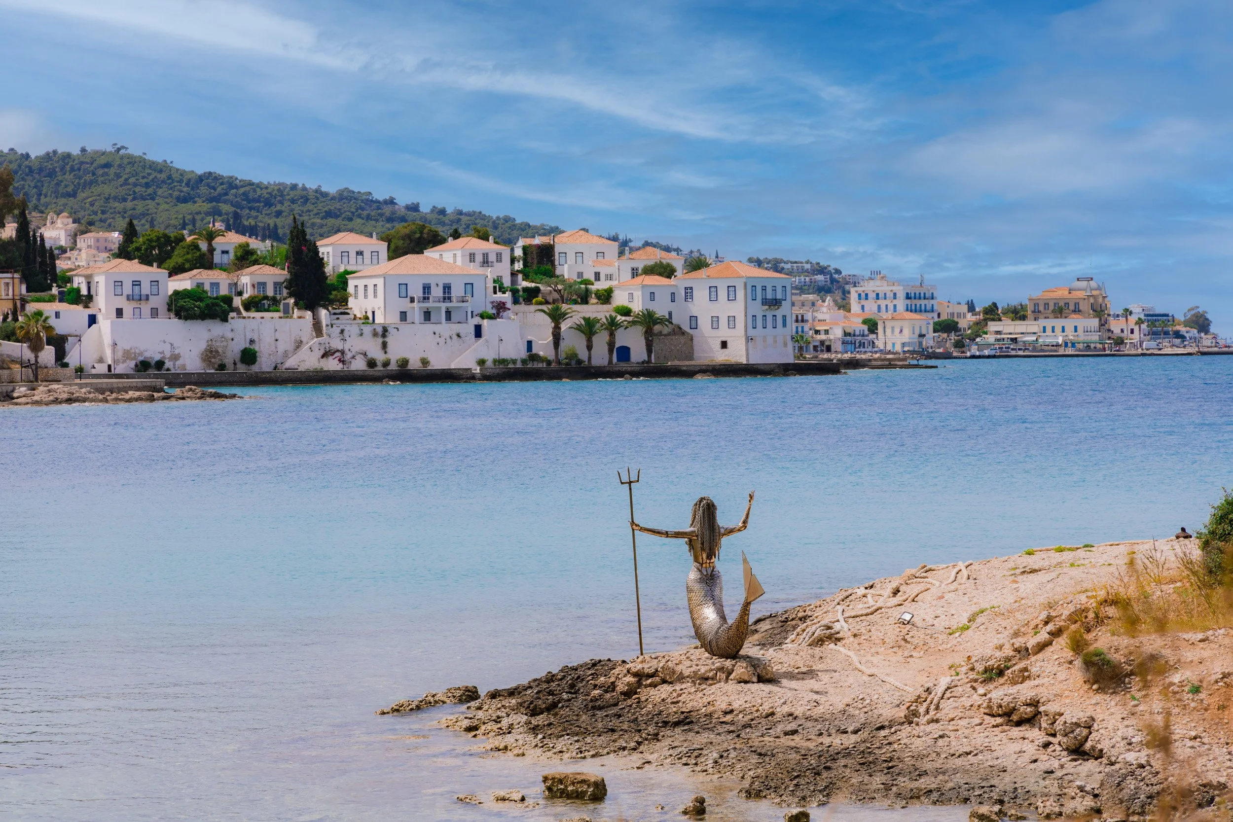 A mermaid sculpture holding a trident and a flag, sitting on a rocky shoreline facing a blue water body with a coastal town in the background, under a partly cloudy sky.