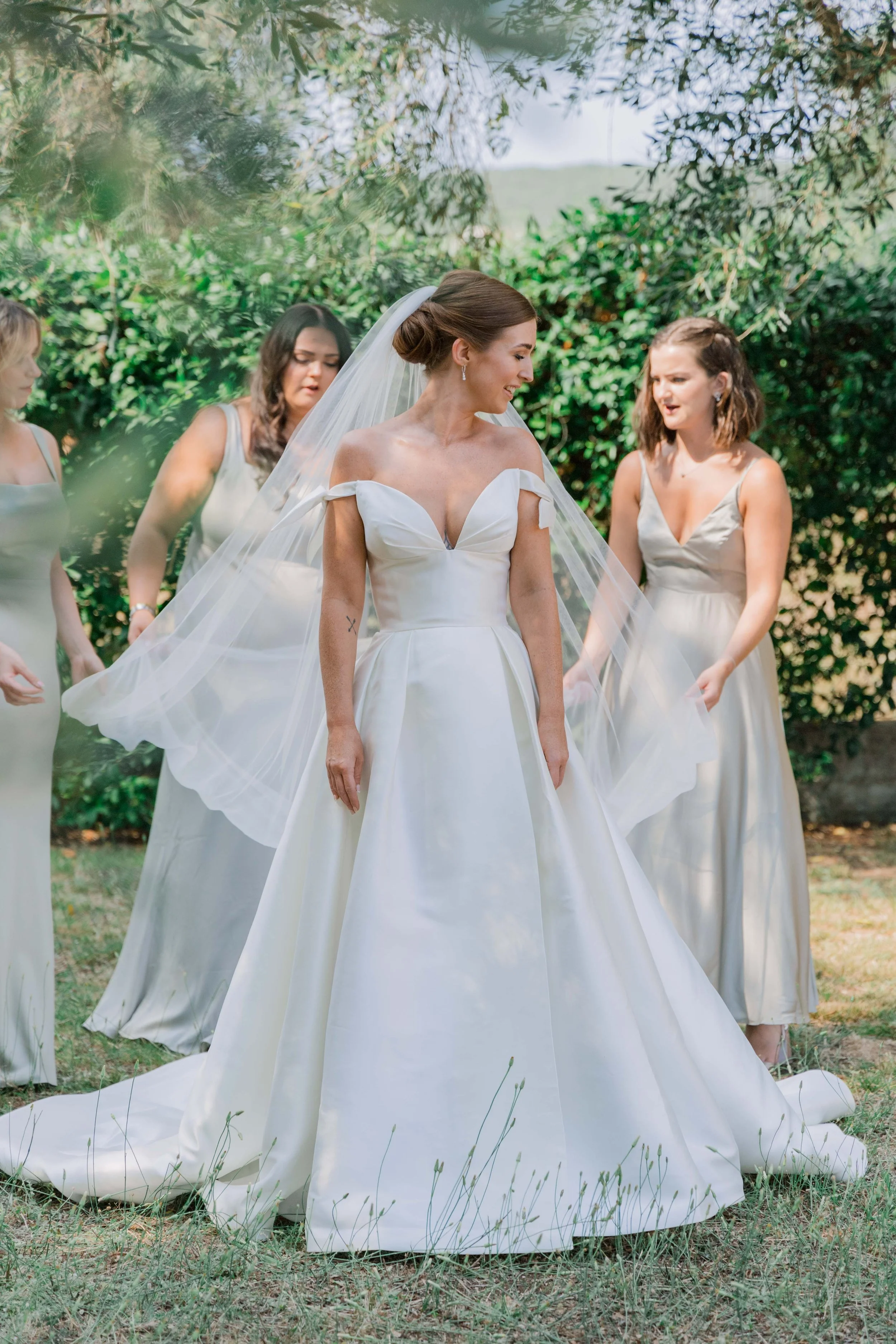 Bride in white wedding gown with off-the-shoulder design, surrounded by her bridesmaids in light-colored dresses, outdoors with greenery in the background.