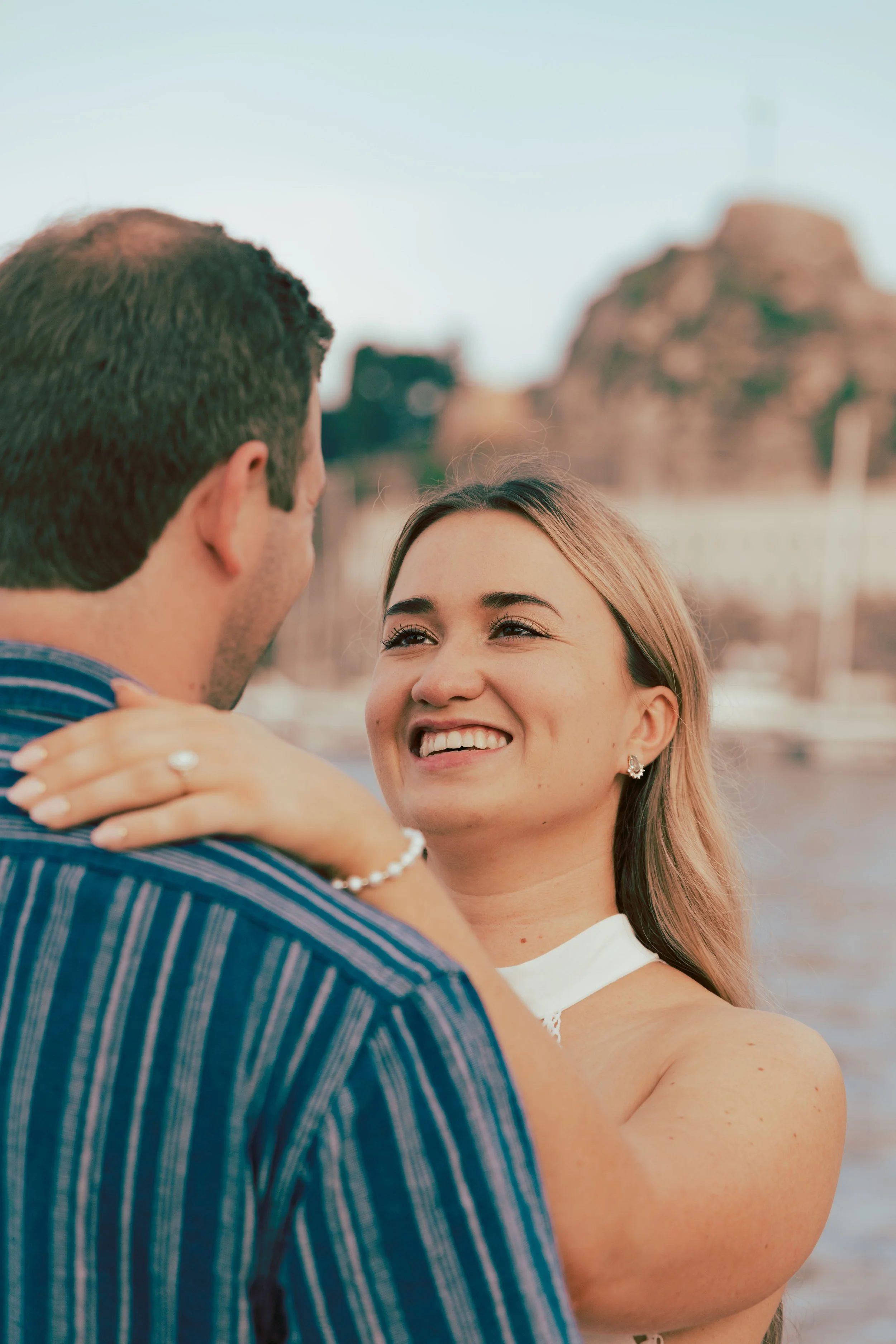 A happy woman with blonde hair smiling at a man while holding him close, with a body of water and rocky cliffs in the background.
