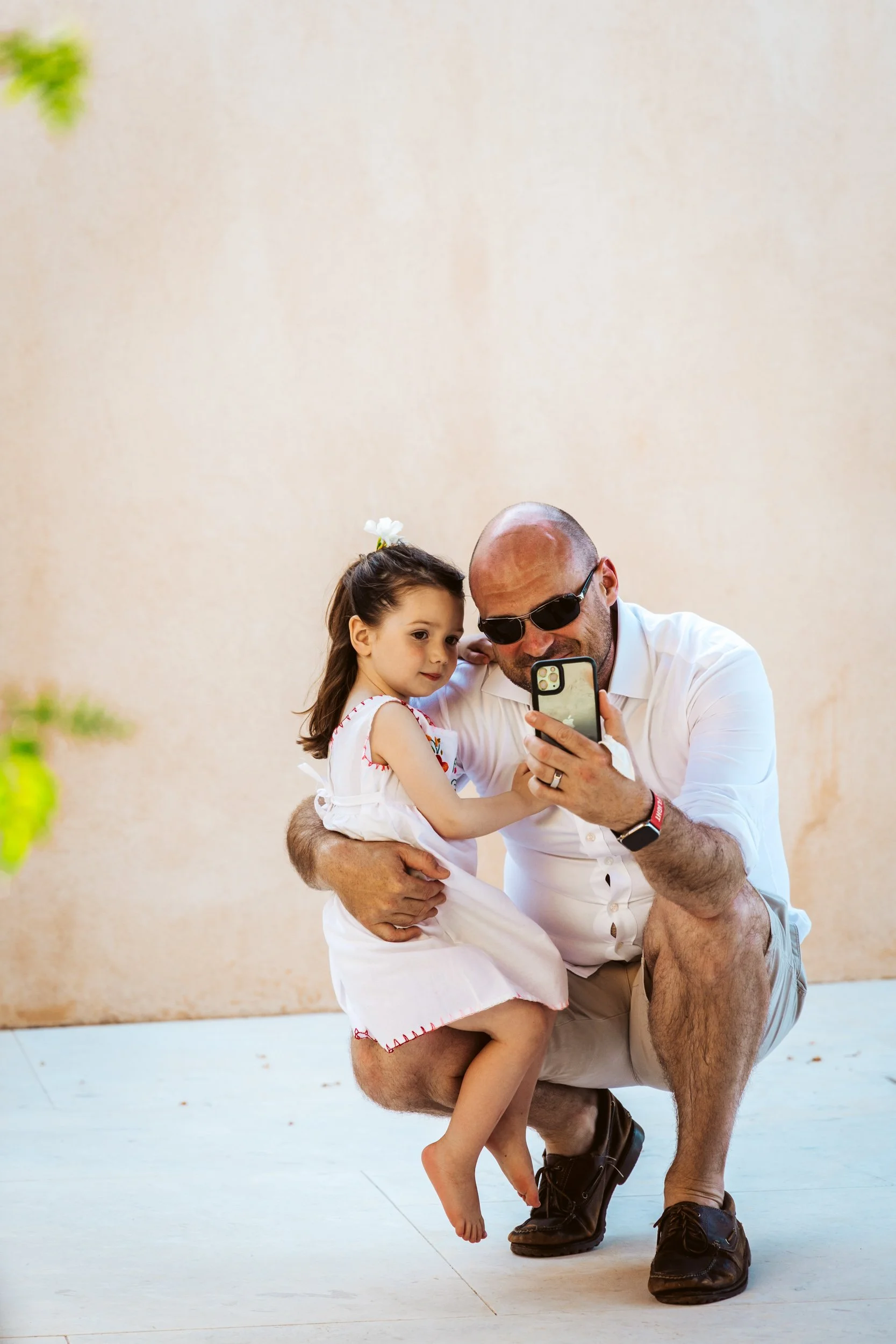 A bald man wearing sunglasses and a white shirt is crouching down on one knee, holding a little girl in a white dress, as they both look at a smartphone screen. The girl has dark hair with a white flower in her hair.