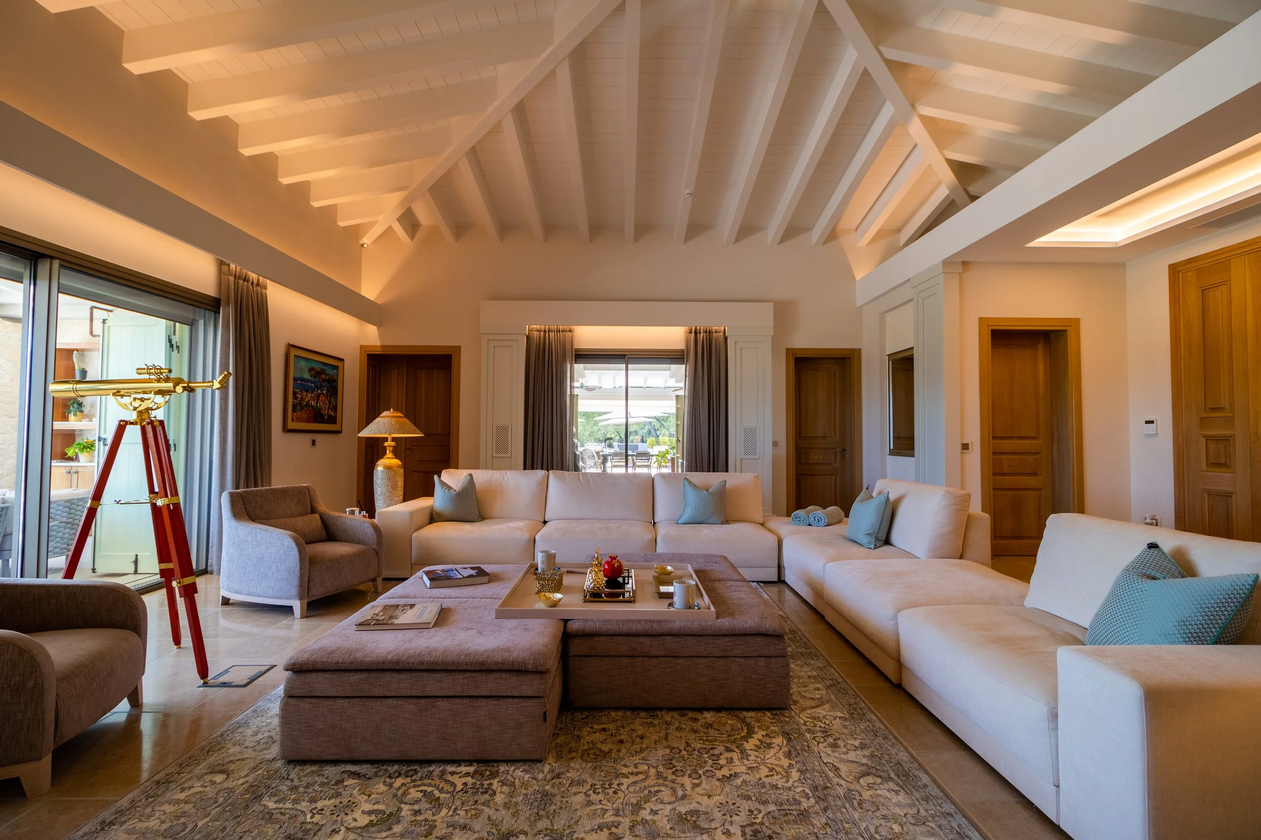 A spacious living room with high white vaulted ceiling, large beige sectional sofa with blue pillows, a patterned rug, a small armchair and a padded ottoman with a tray of decorative items on it. There are two sliding glass doors, curtains, a telesco