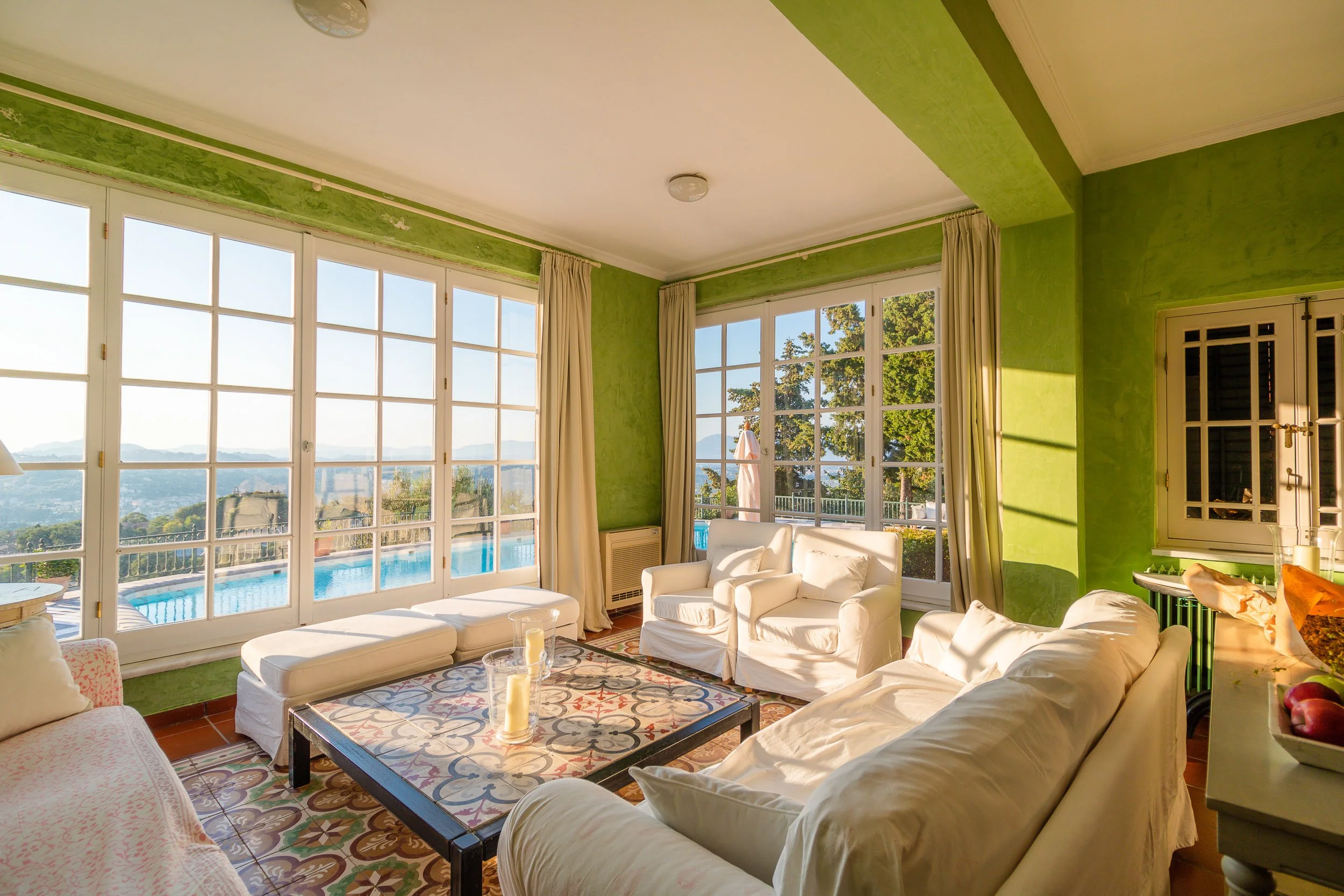 Bright living room with large windows, white sofas, and a colorful tiled coffee table overlooking a pool and scenic landscape.