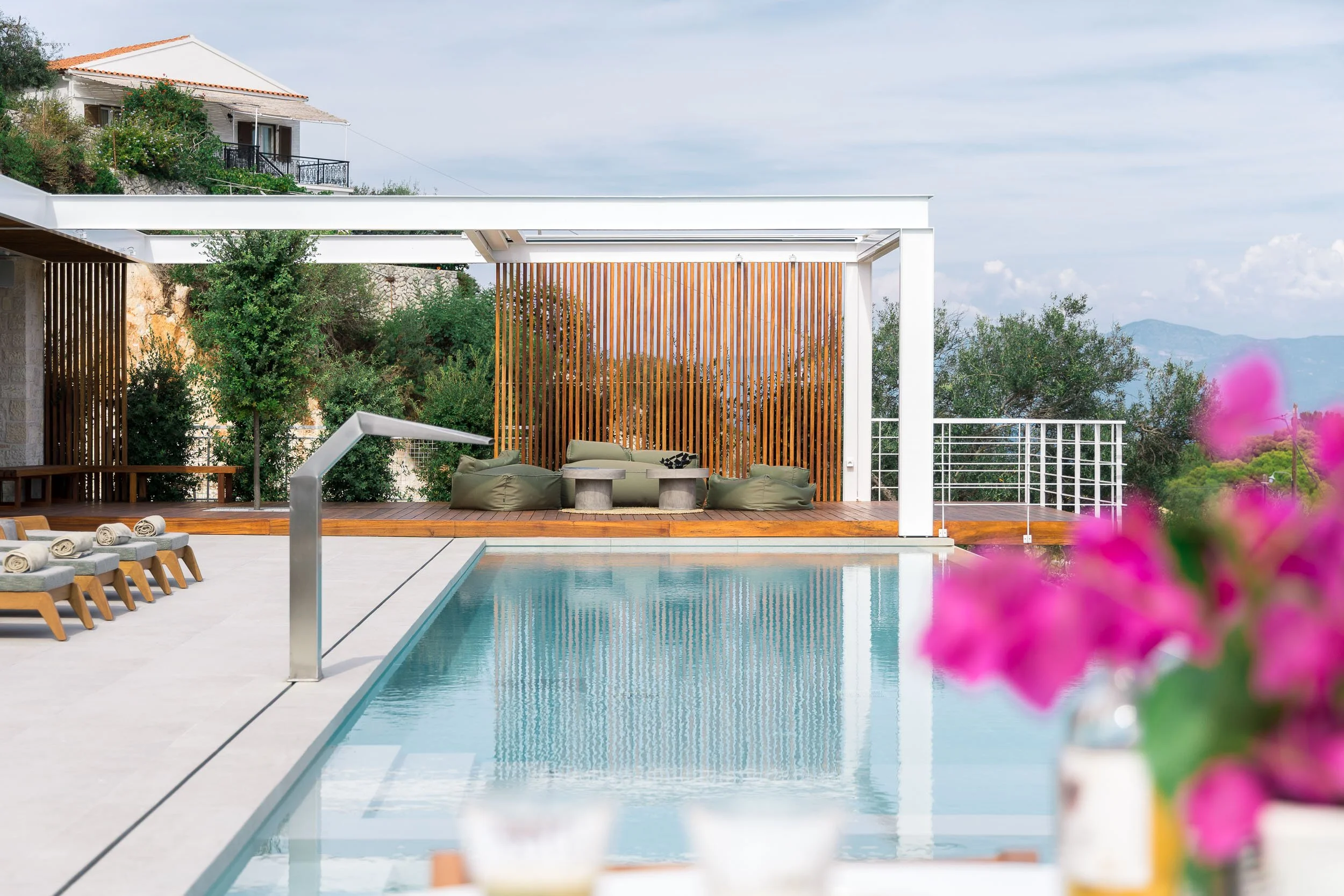 Modern outdoor terrace with a swimming pool, lounge chairs, a shaded seating area with bean bag chairs, and a scenic view of trees and mountains in the background.