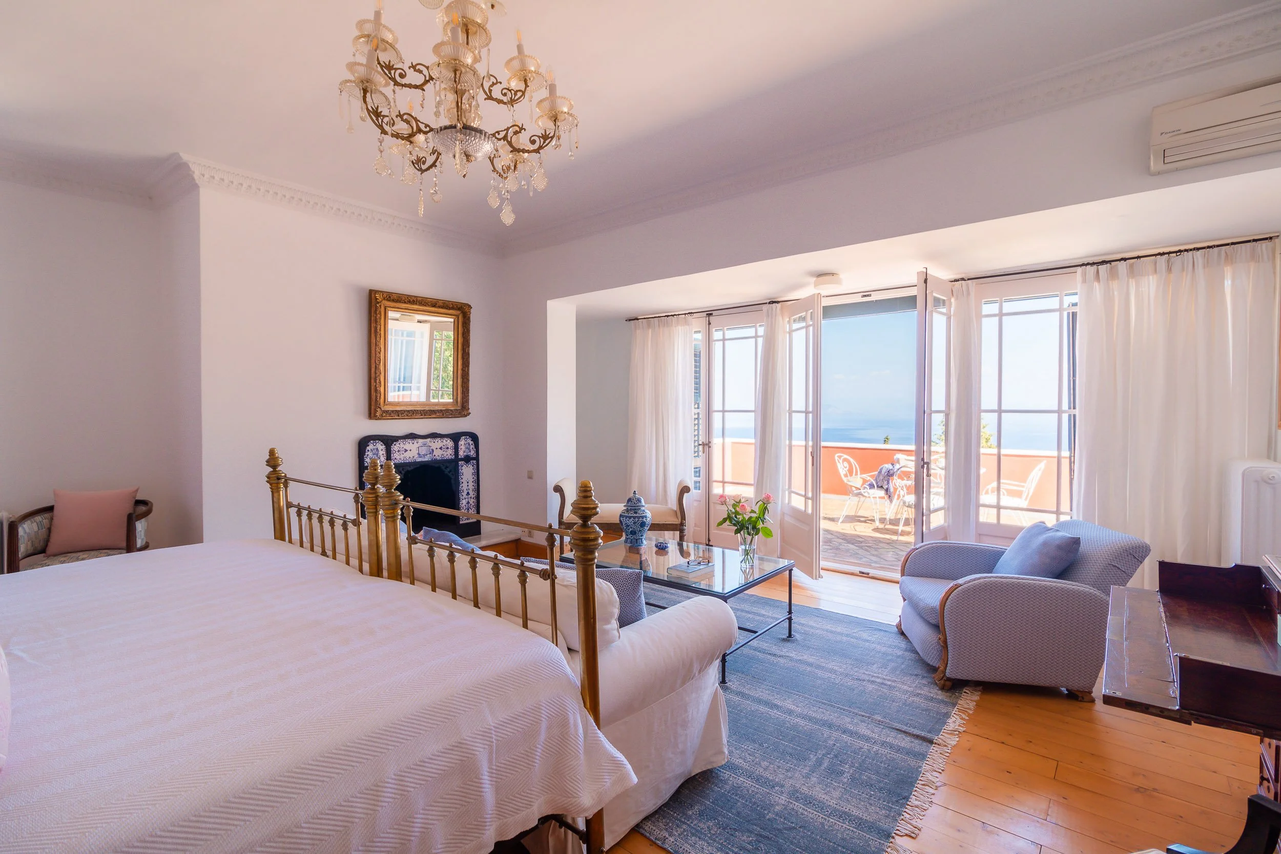 A bright hotel bedroom with a large bed, sitting area, and a balcony with outdoor furniture. The room has white walls, wooden flooring, and large windows with white curtains, overlooking the sea.