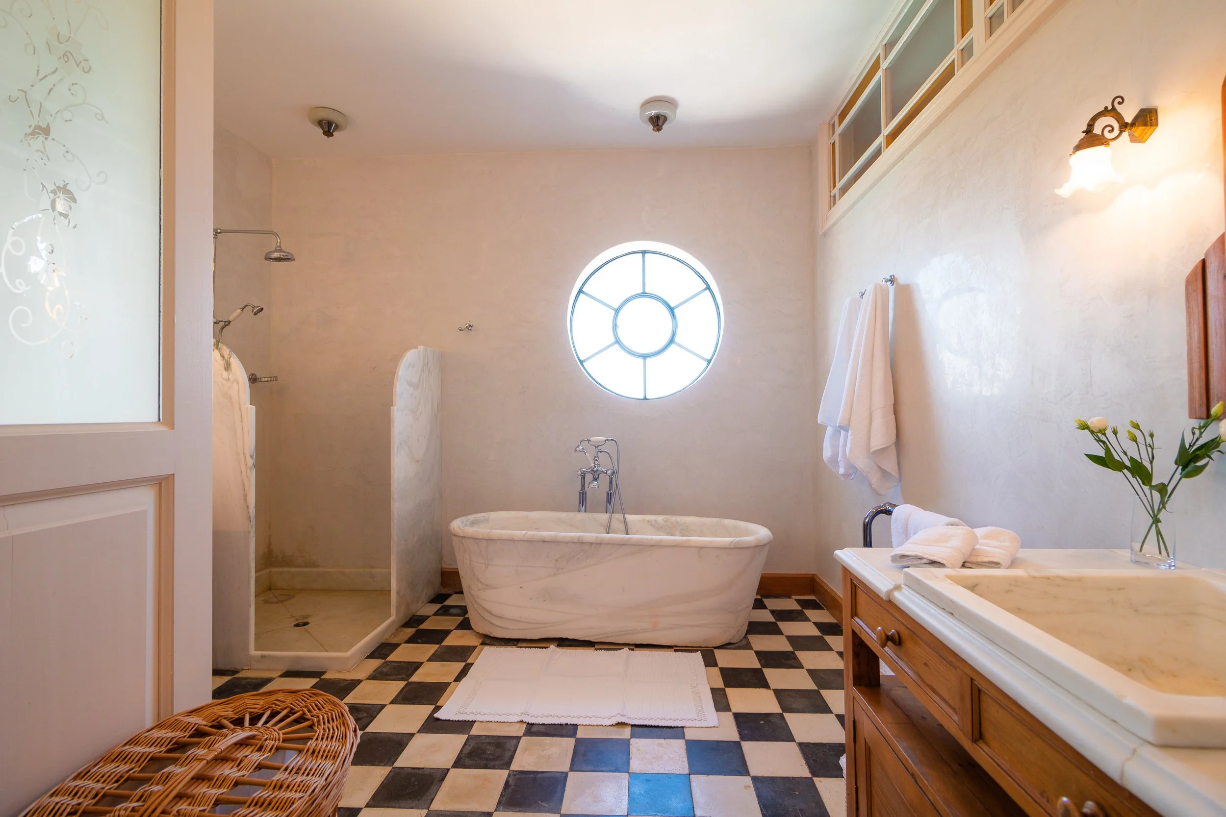 Bathroom with a marble bathtub, a walk-in shower, a window, wooden vanity with a vase of flowers, towels, and checkered black and white floor tiles.