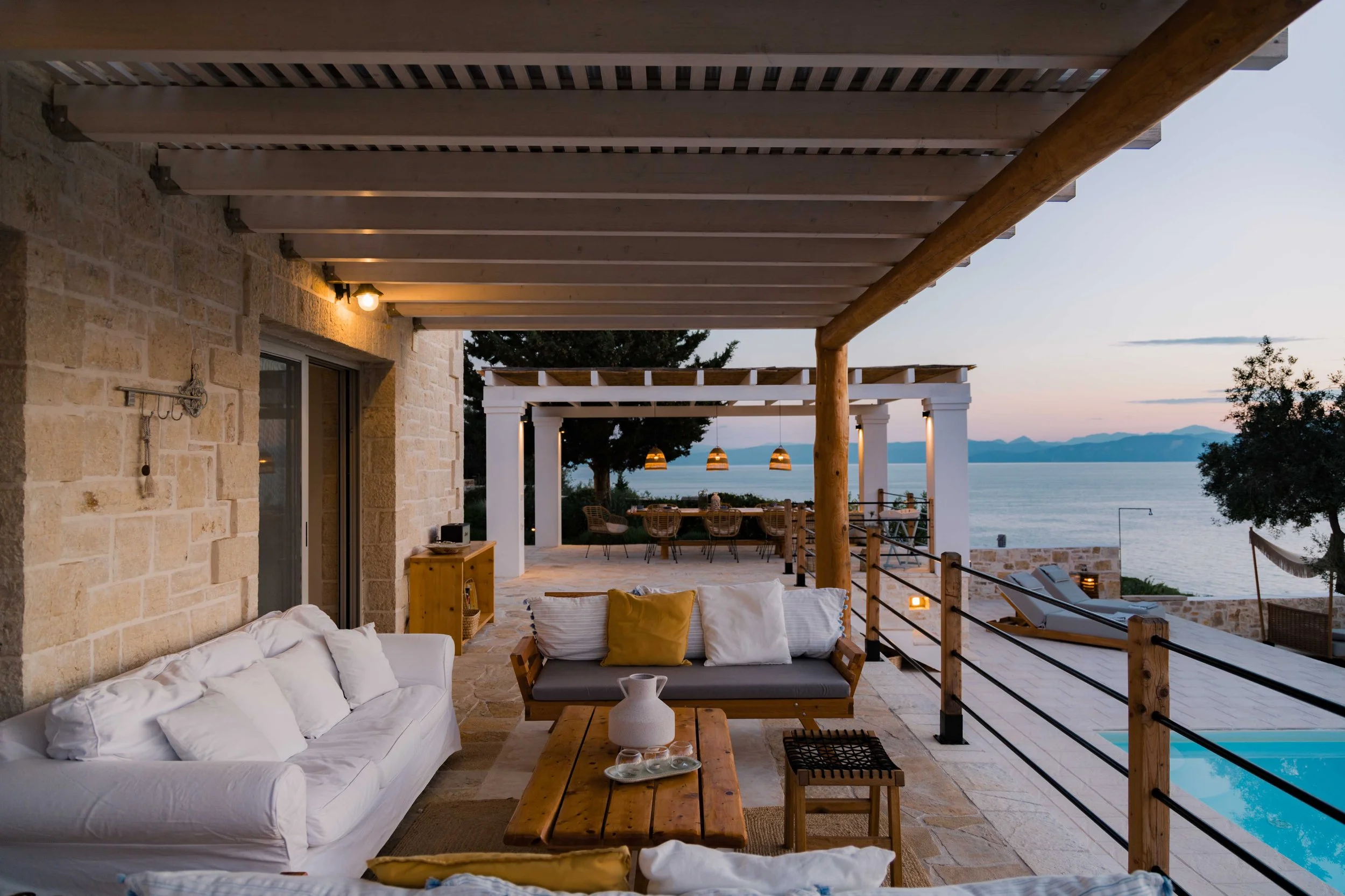Outdoor patio with seating area facing a body of water at sunset. Features a white sofa, a wooden bench with cushions, and a dining area under a pergola with hanging lights. Views of mountains and a clear sky in the background.