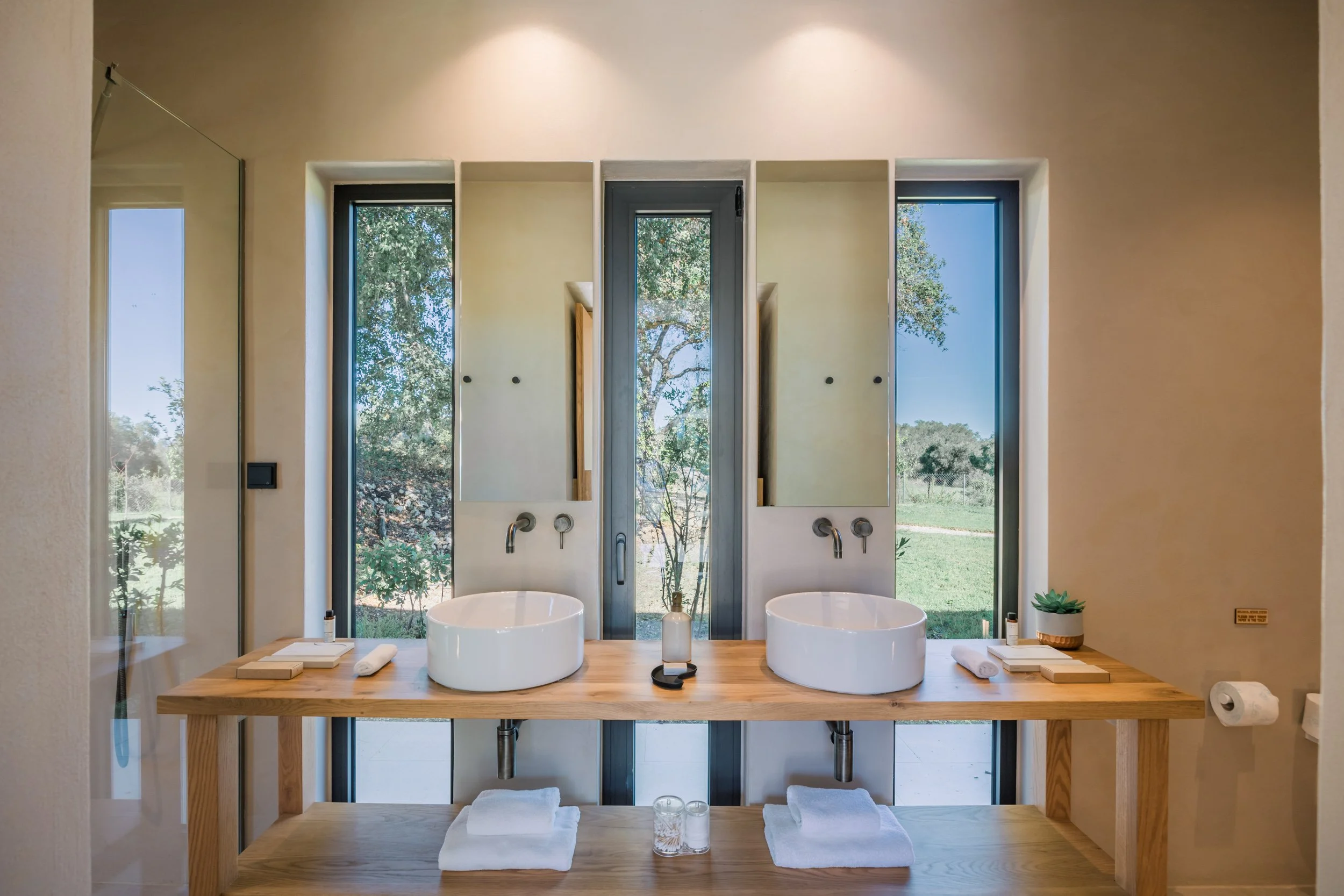 Modern bathroom with two white vessel sinks on a wooden vanity, large windows with views of trees and grass, minimalist decor, and natural light.