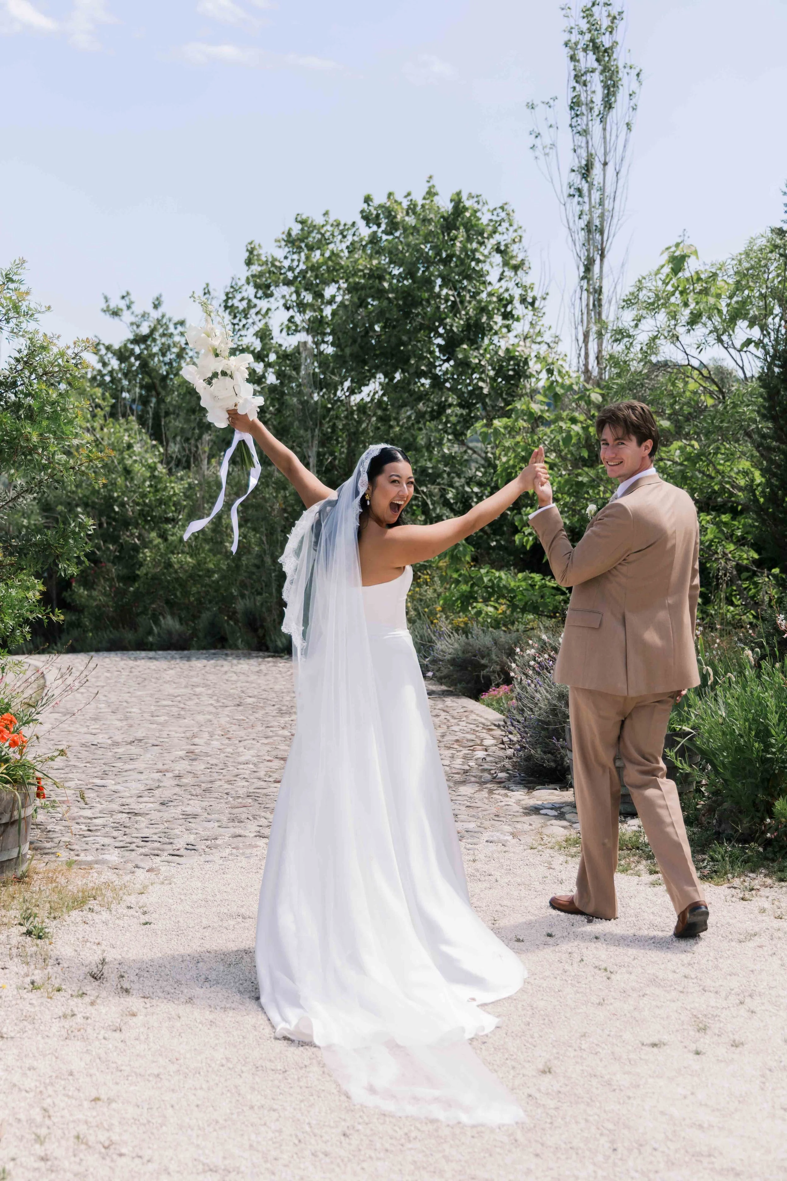 A bride and groom celebrating outdoors during their wedding, with the bride waving a bouquet and the groom holding her hand, surrounded by green trees and a sunny sky.