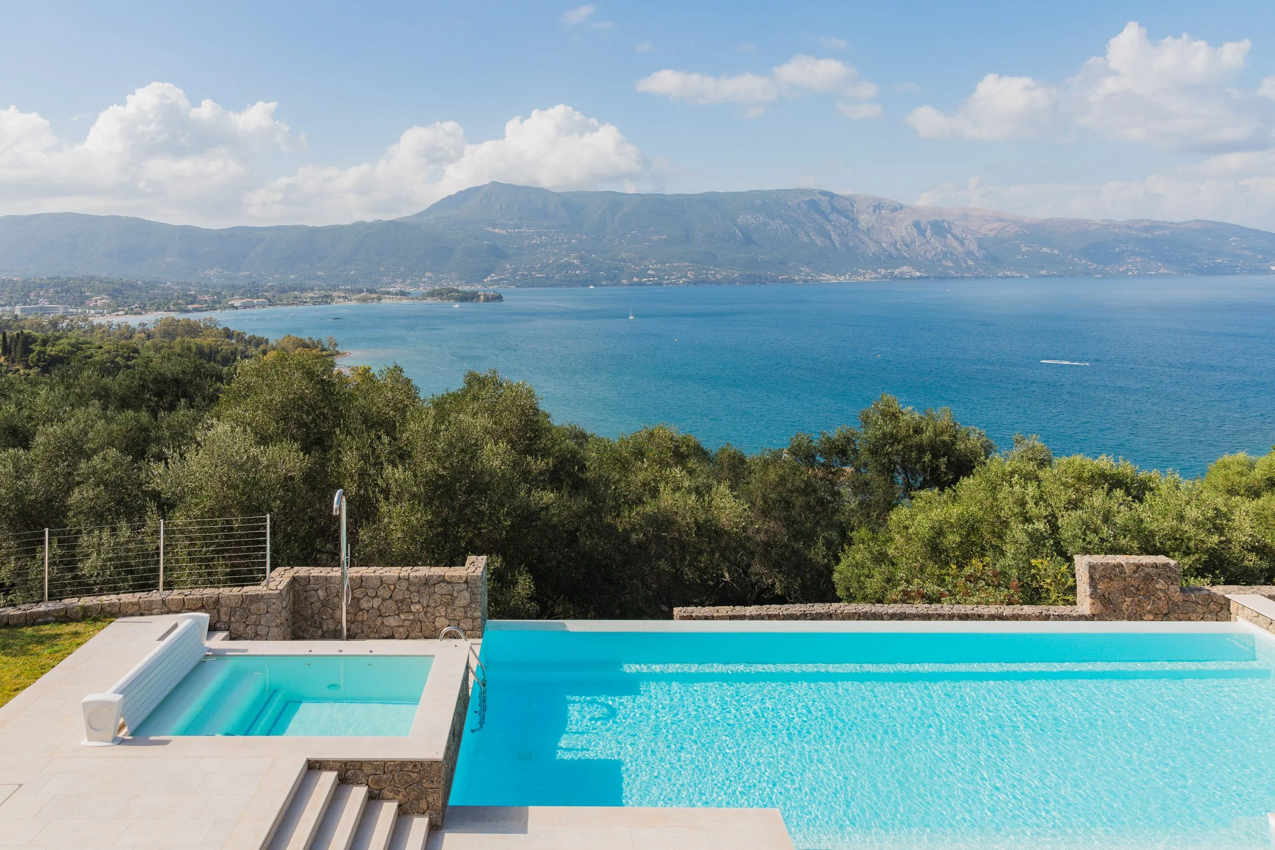 Infinity pool overlooking a lush green landscape and a large body of water with mountains in the background under a partly cloudy sky.