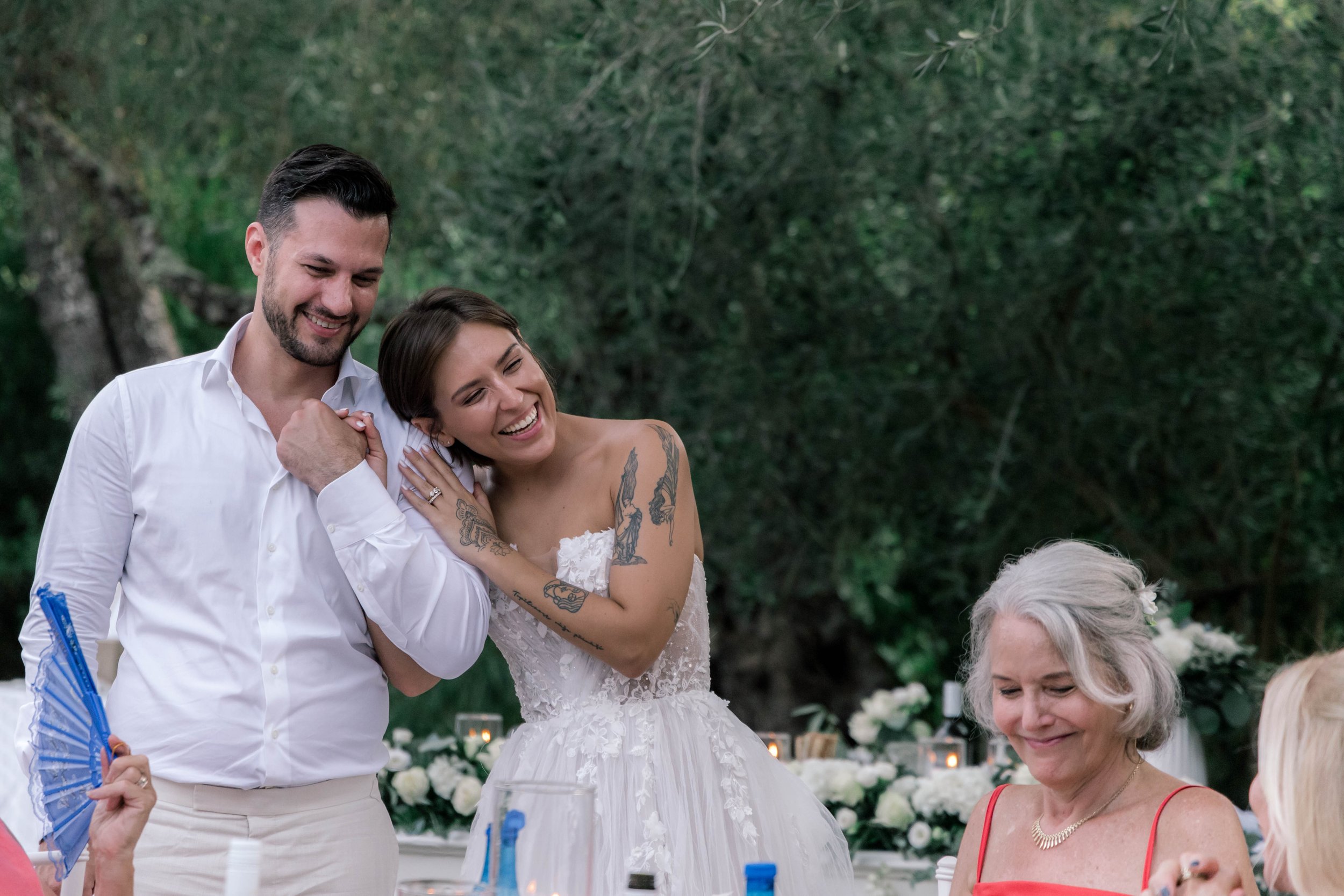 A bride and groom smiling and embracing each other at a wedding reception outdoors, surrounded by guests and floral decorations.