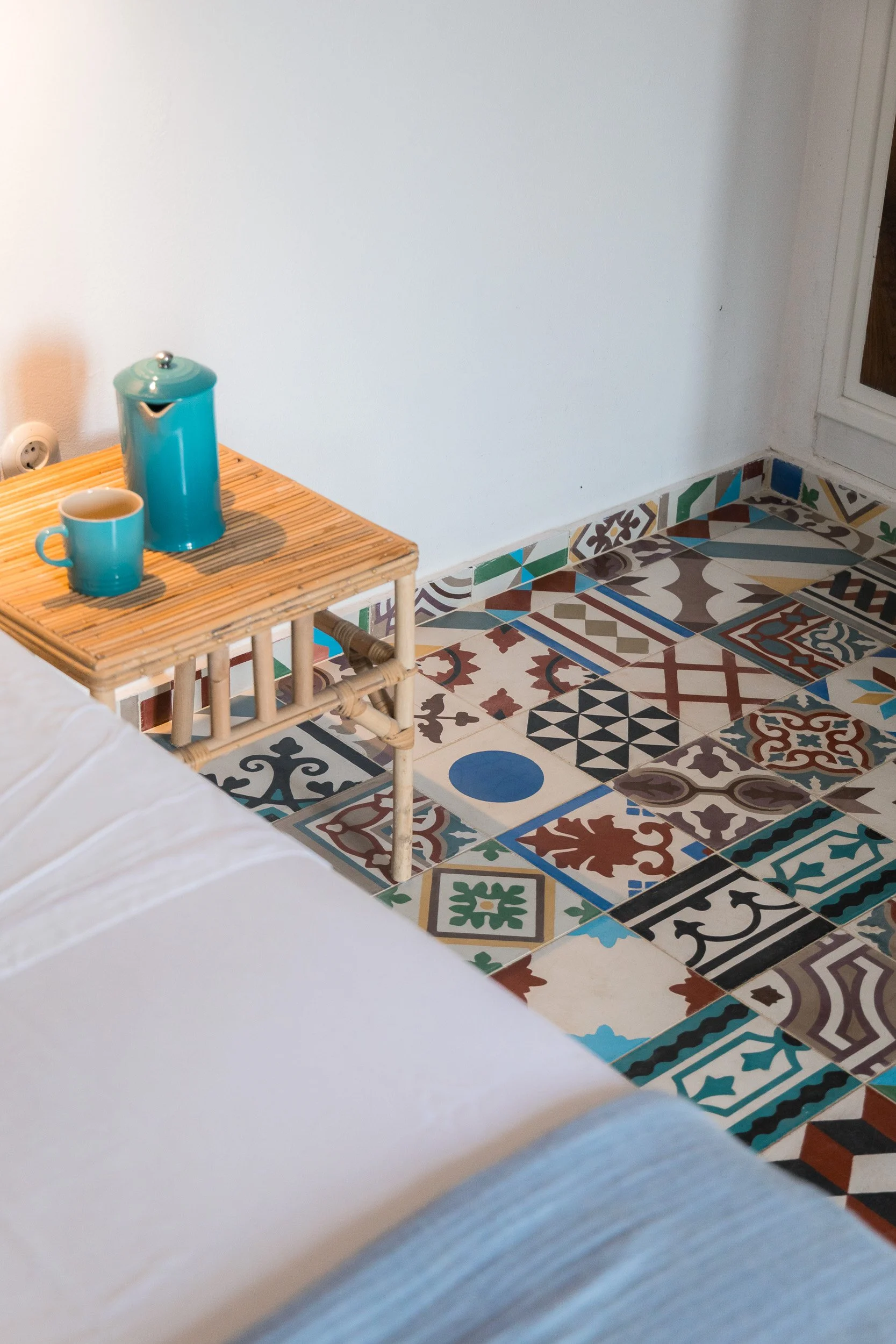 A small wooden side table with a turquoise coffee mug and a matching turquoise coffee canister, placed next to a white sofa, in a room with patterned colorful tile flooring.