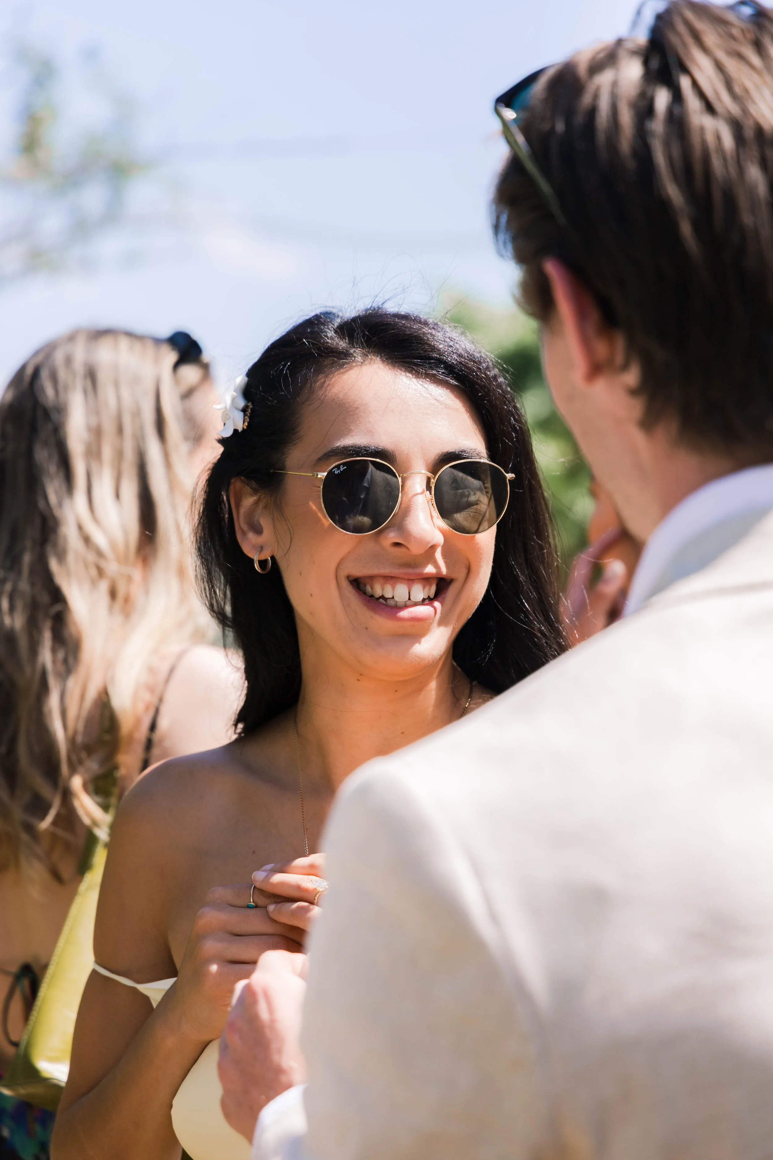 A woman wearing sunglasses and a white off-the-shoulder top, smiling and engaging in a conversation outdoors with a man in a white jacket, surrounded by other people on a sunny day.