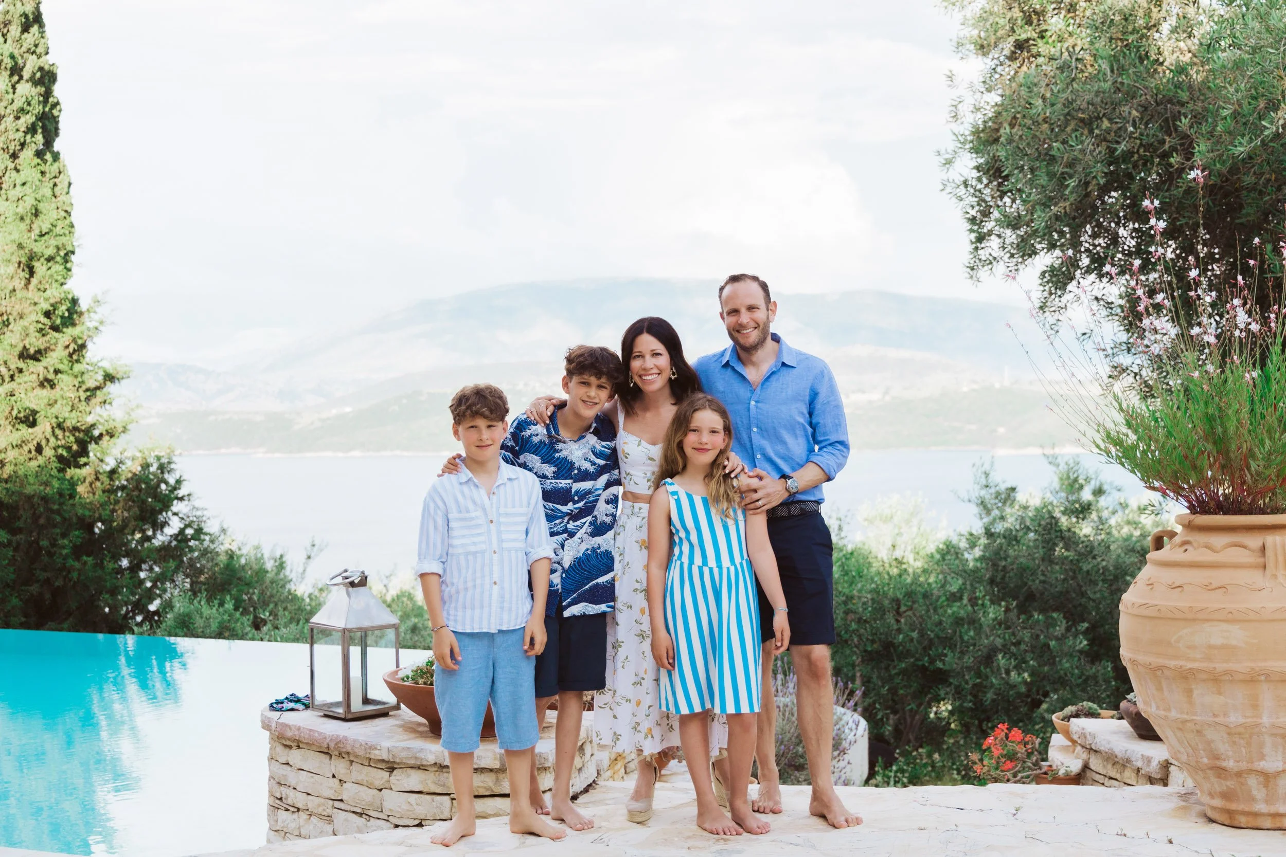 A family of five posing outdoors near a pool, with mountains and a lake in the background, surrounded by greenery and large potted plants, all dressed in summer outfits.