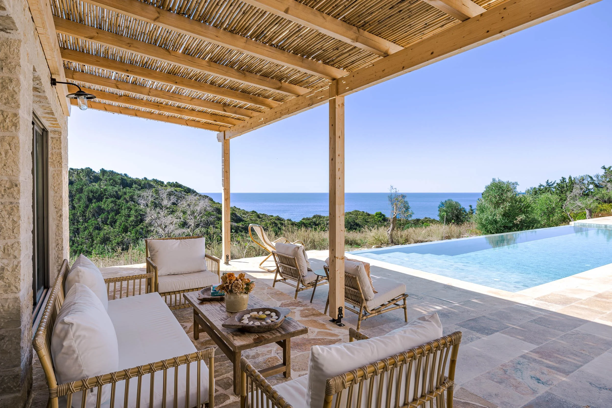Outdoor patio area with wicker furniture, a wooden table with a floral centerpiece, two lounge chairs, and an infinity pool overlooking a landscape of trees and the ocean under a clear blue sky.