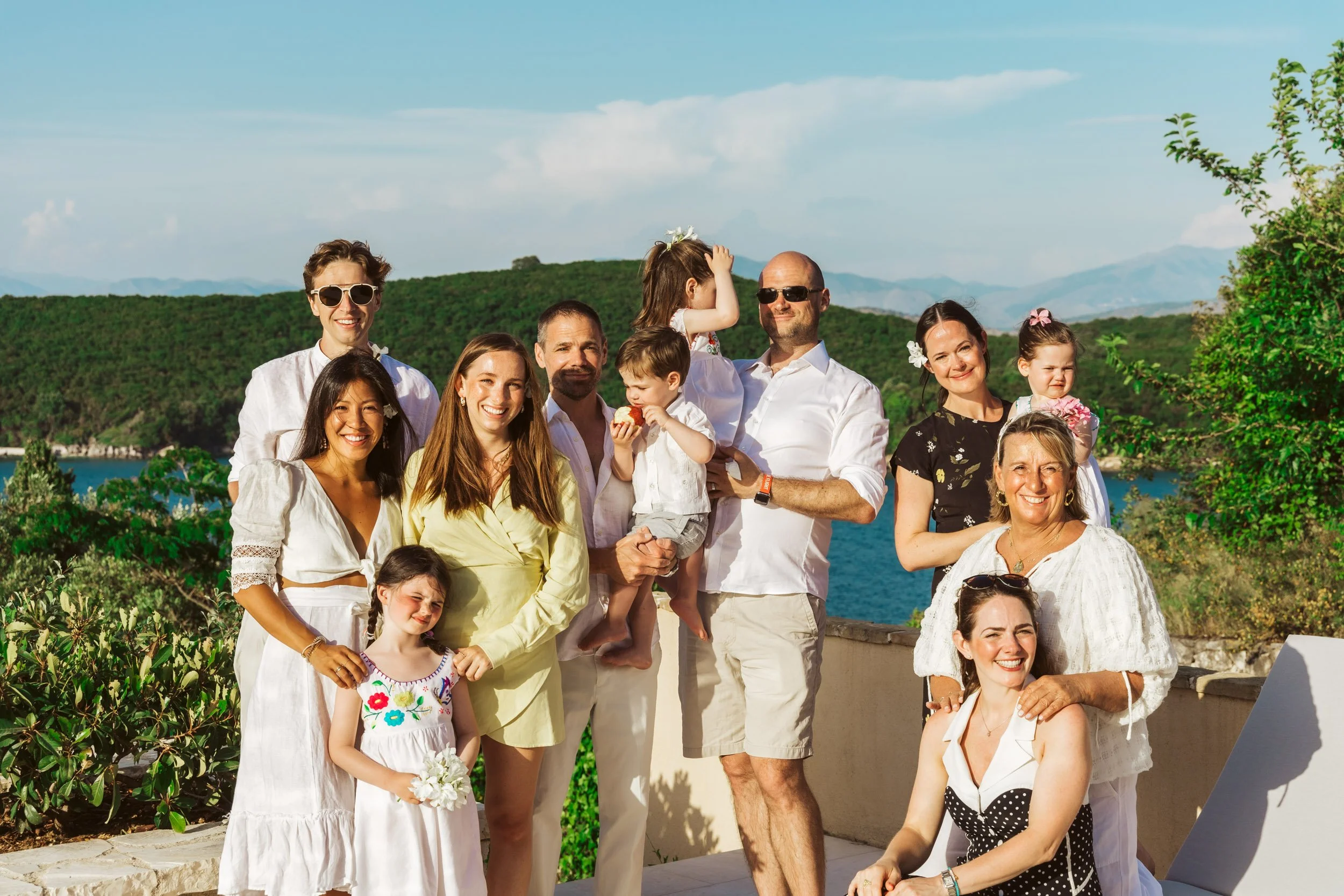 A group of people, adults and children, smiling and gathered outdoors on a sunny day with a mountain and water in the background.